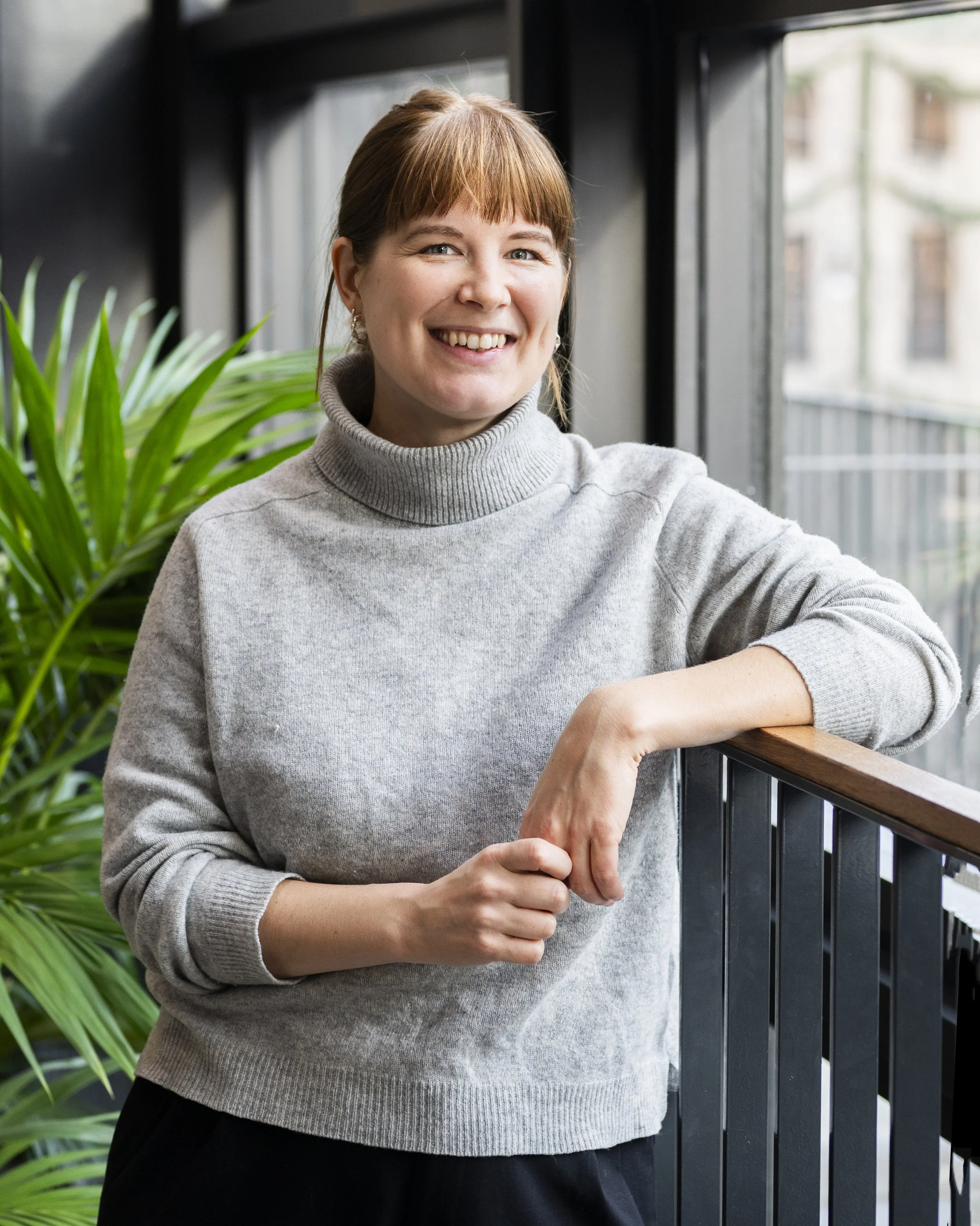 Portrait image of Julia, smiling and looking into the camera. She wears a gray sweater and there's a green palm tree in the background.