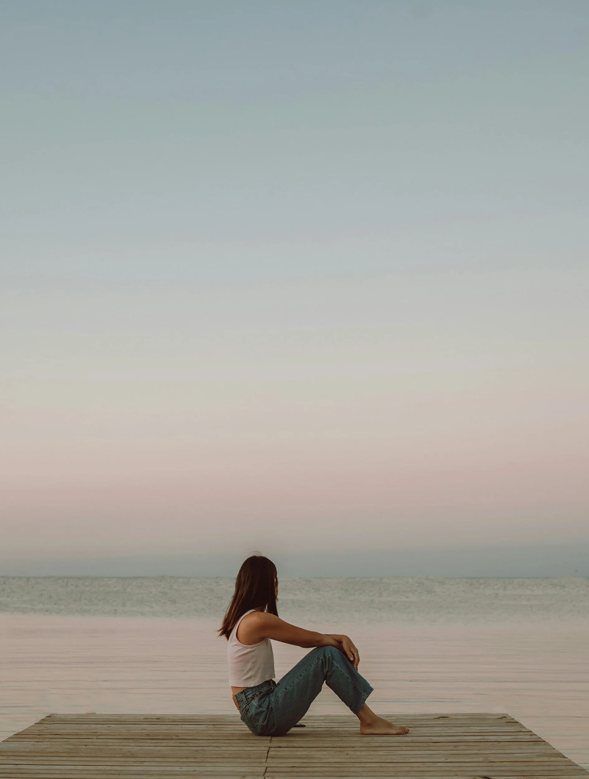 A woman wearing a white tank top and jeans is sitting on a wooden dock at the beach, looking out over the water with a calm expression during sunset or dusk.