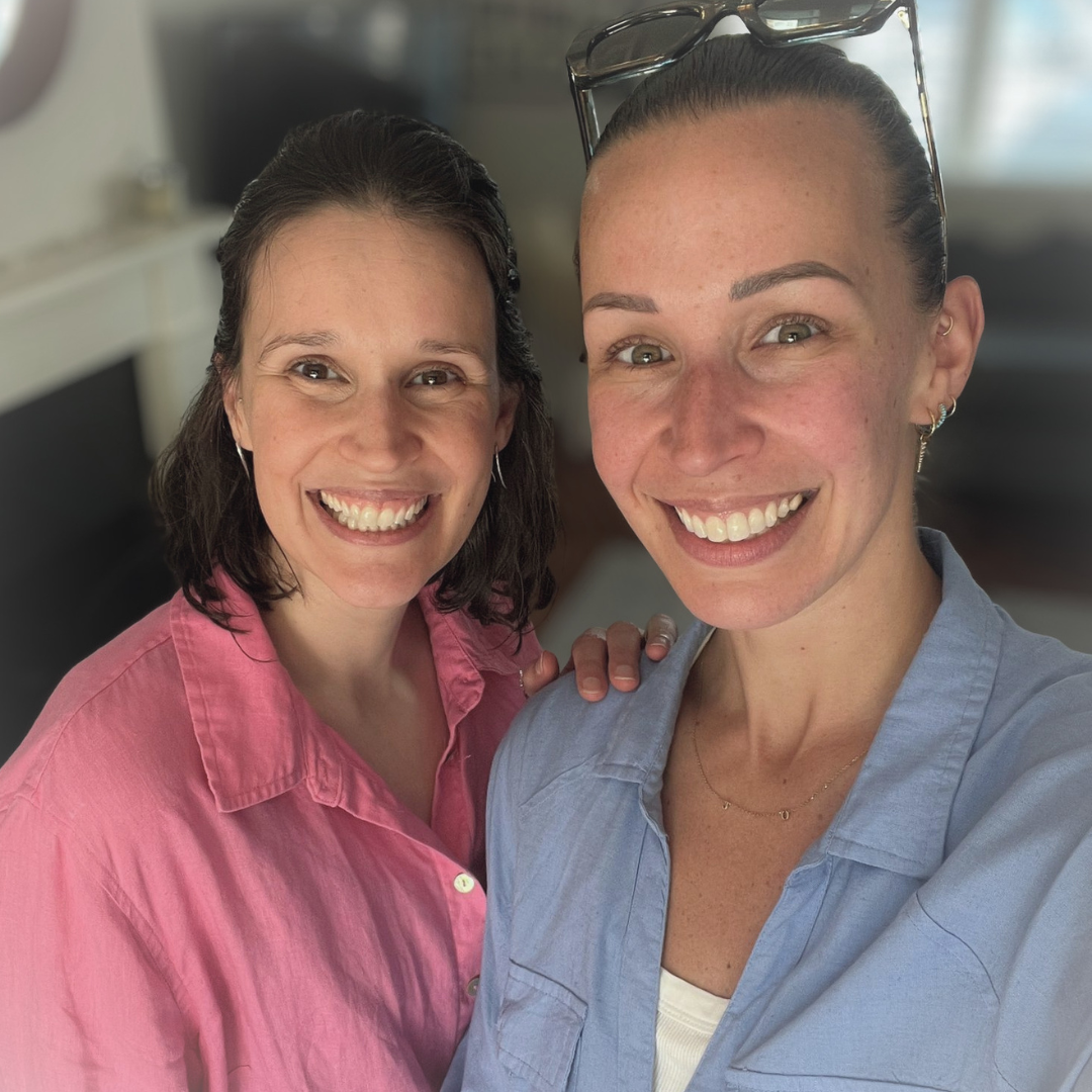 Two women smiling, one in a pink shirt and the other in a blue shirt, taking a close-up selfie indoors.
