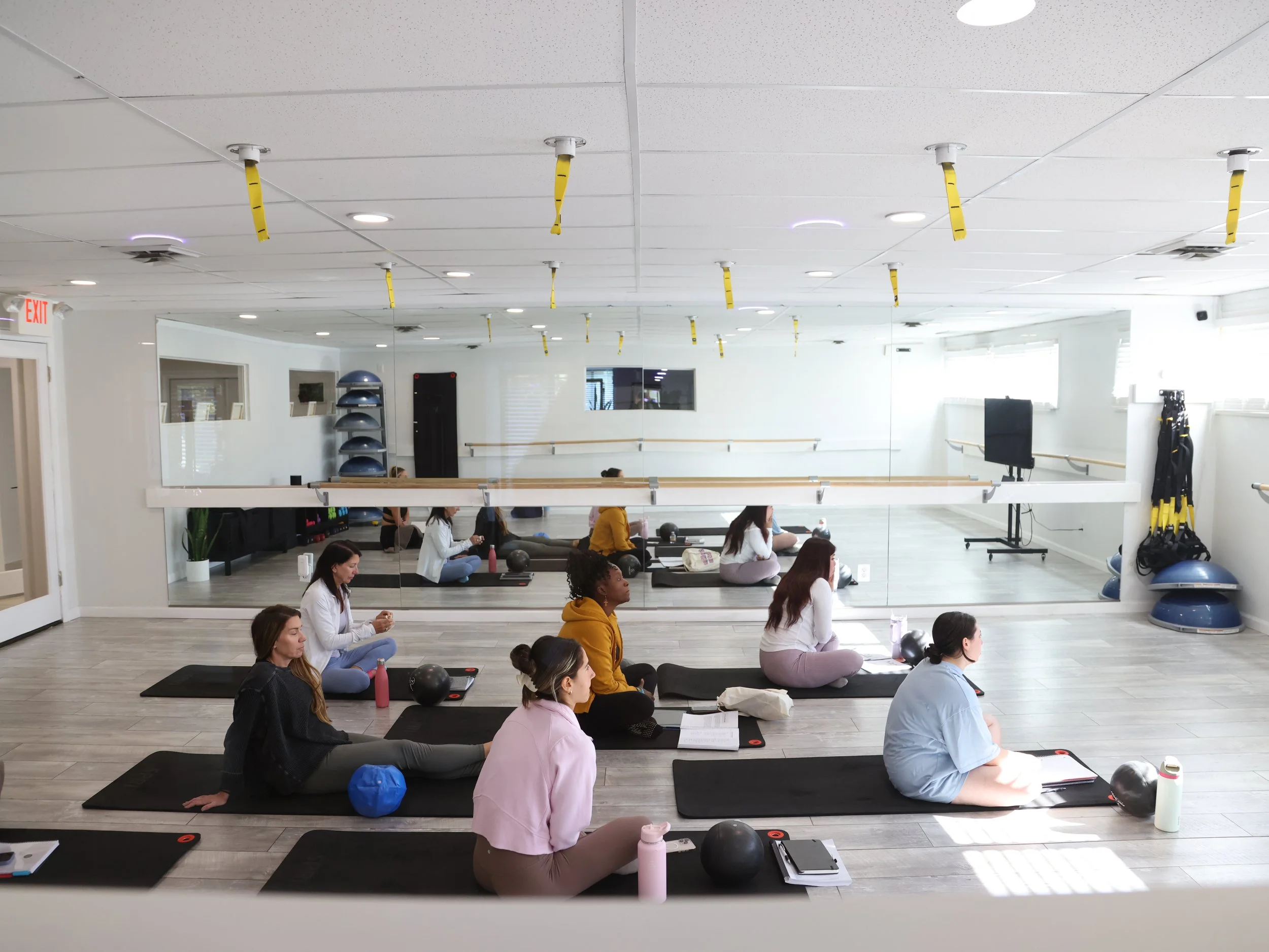 Three women practicing yoga indoors on a black mat, with three potted plants in the background.