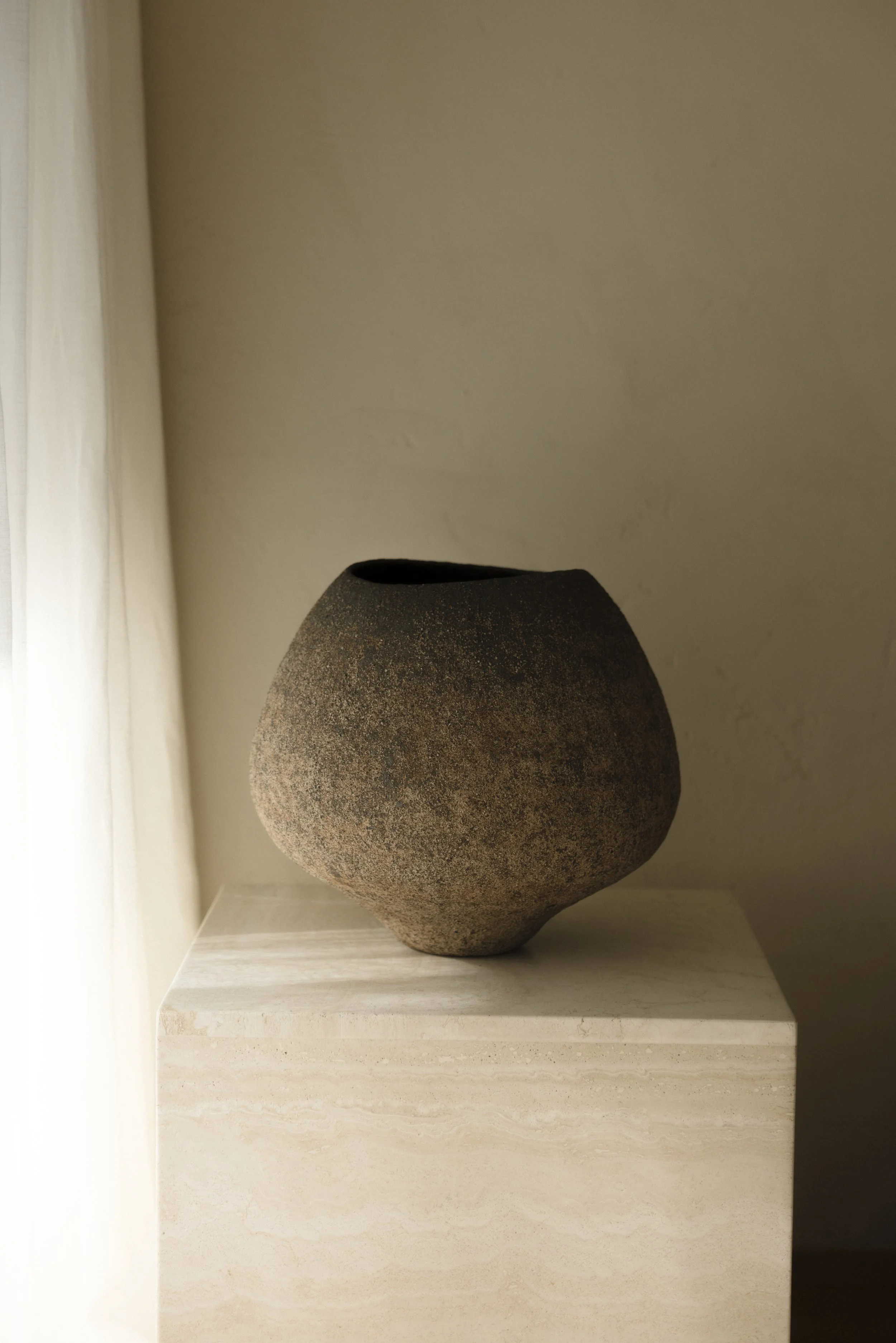 A dark, textured ceramic vessel by artist Hiroshi Aoki displayed on a minimalist travertine stone plinth against a lime-wash wall.