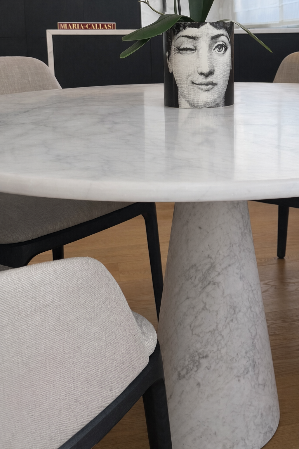A marble-topped round table with a cylindrical marble base, surrounded by beige cushioned chairs with black frames. On the table, there is a black and white portrait of a woman with one eye winking, placed inside a cylindrical container with a plant. In the background, a framed sign reads 'Maria Callasi.'