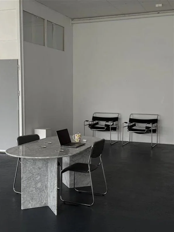 Empty office room with a marble conference table, four black chairs, a laptop, a coffee mug, and two black chairs against the white wall.