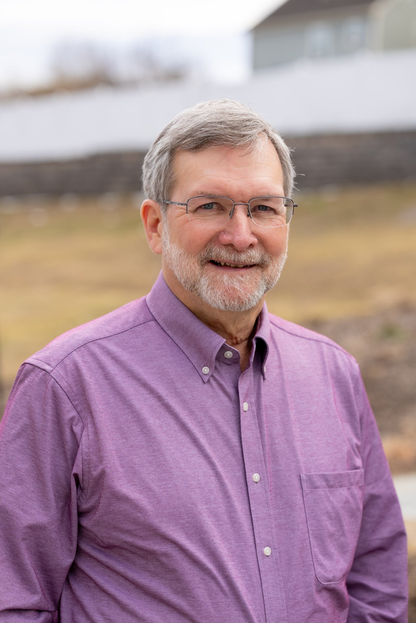 A middle-aged man with gray hair and glasses, wearing a dark pinstripe suit, light blue shirt, and a pink patterned tie, smiling outdoors during sunset.