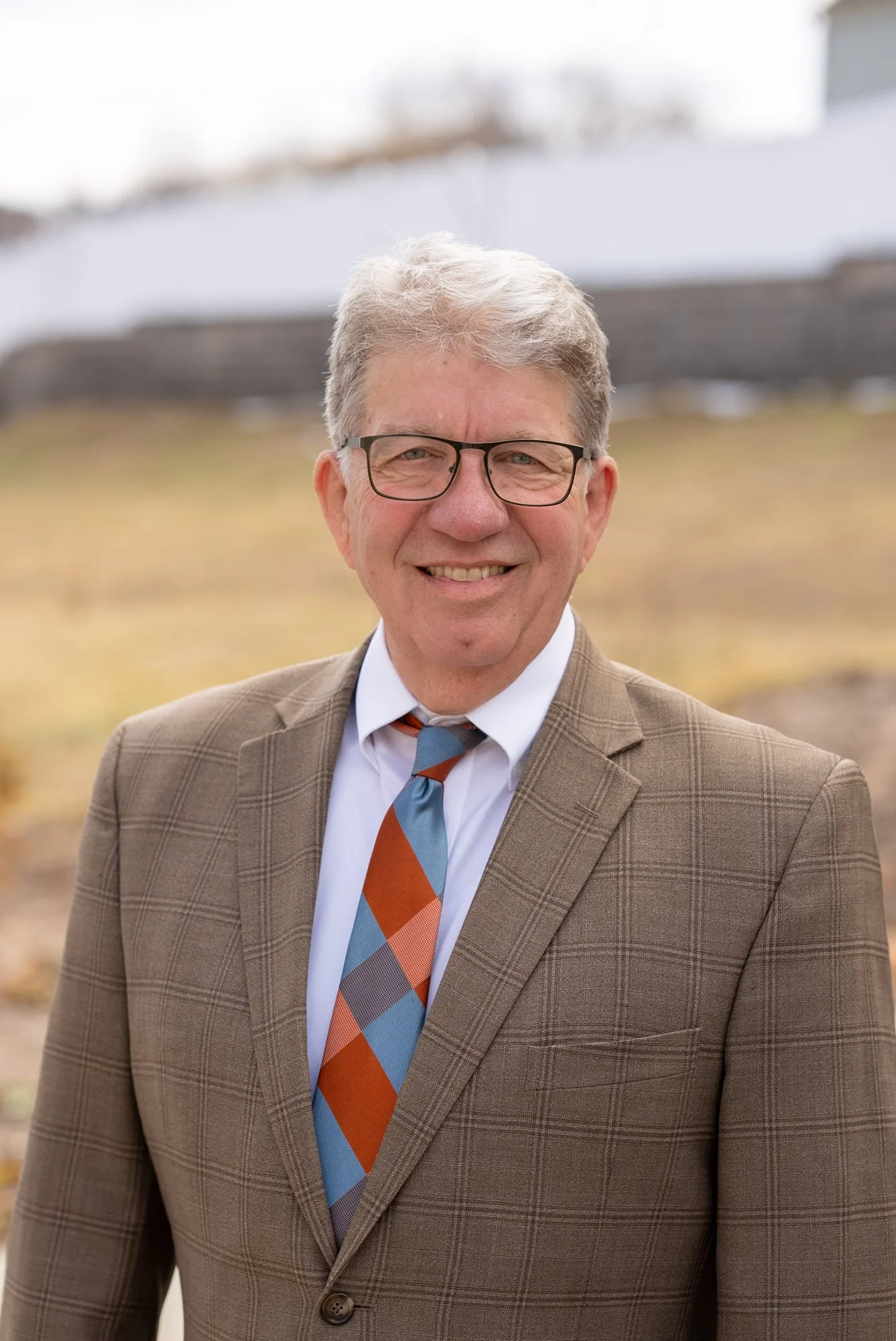 A man in a white shirt, black vest, and glasses smiling in an indoor setting with framed pictures and a wooden wheel in the background.