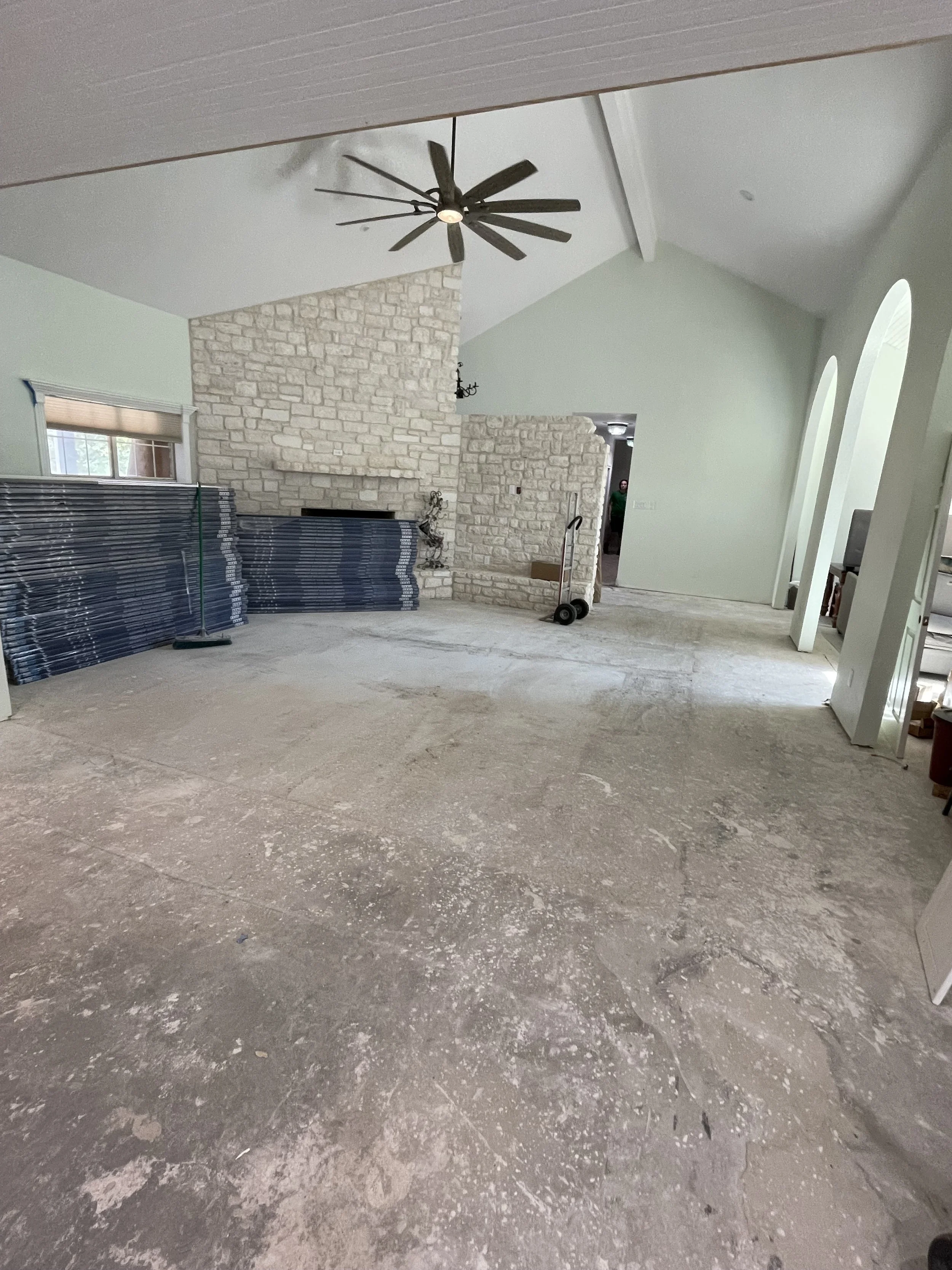 Living room under renovation with exposed concrete floor, a stone fireplace, a ceiling fan, and light green walls.