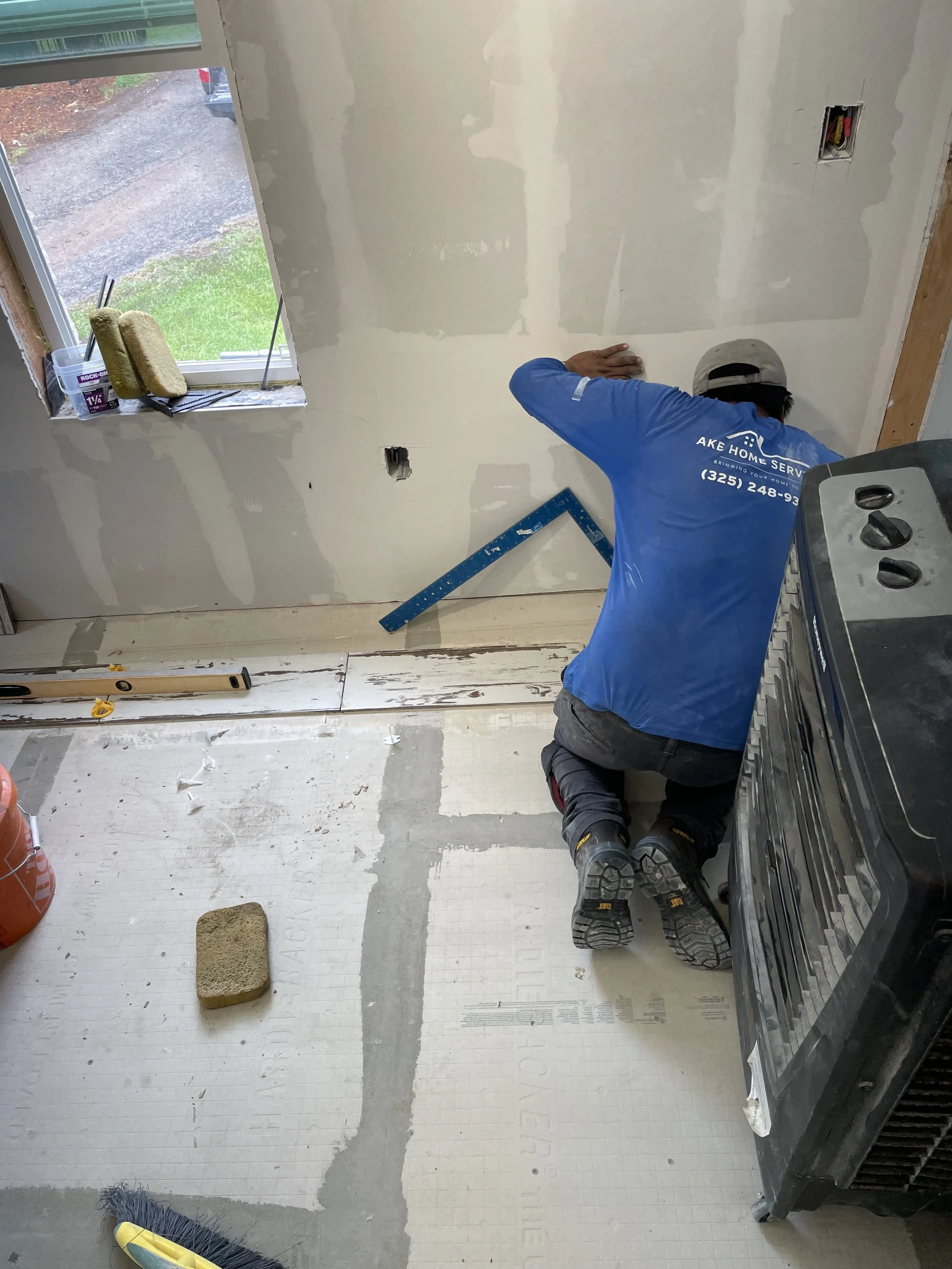 A construction worker kneeling on the floor of a room under renovation, working on drywall near a window, with tools and supplies scattered around.