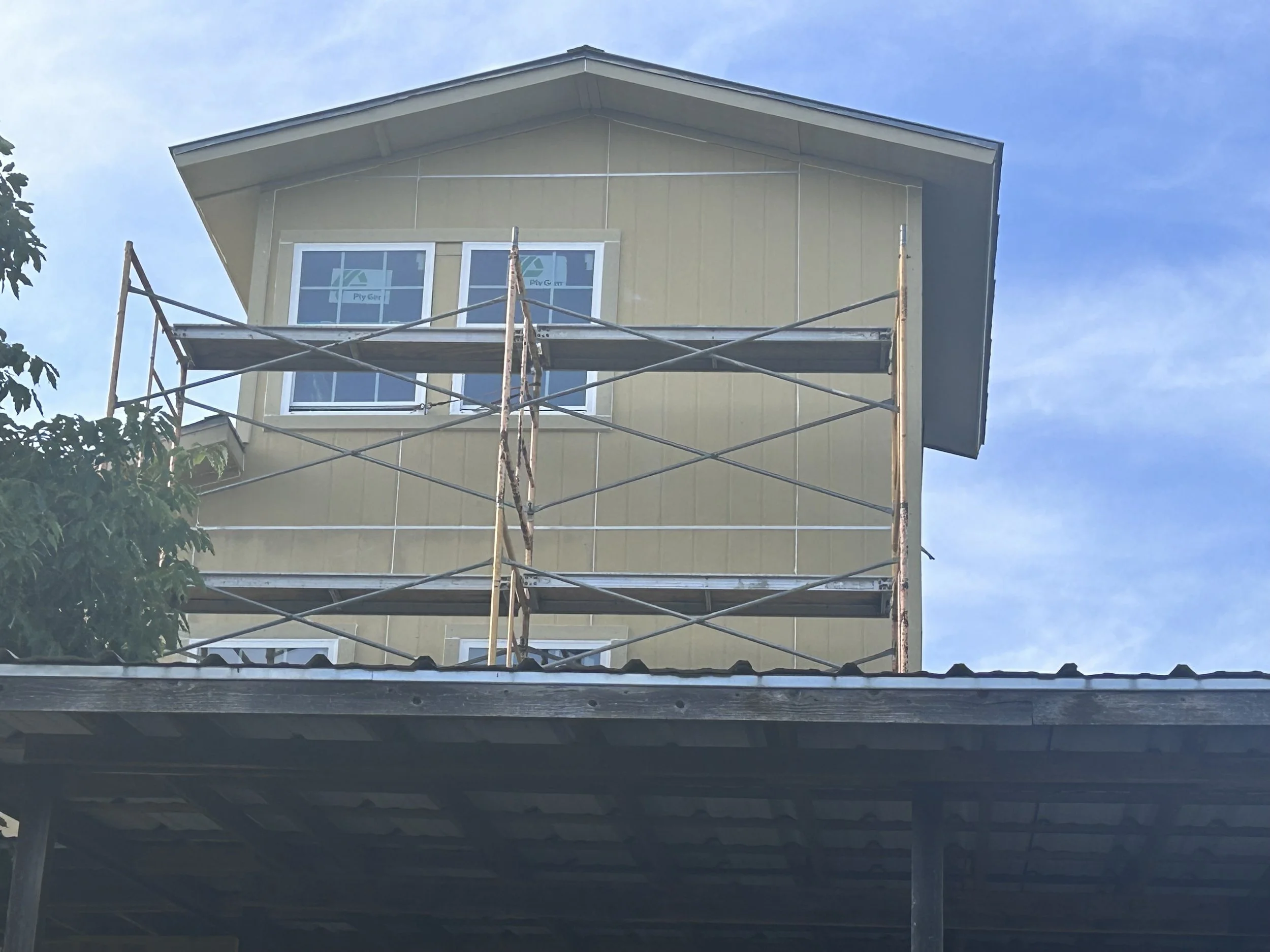 A house under construction with beige siding, two window panes with white frames, and scaffolding in front of the upper windows. A leafy tree is partially visible on the left side, and the sky is partly cloudy.