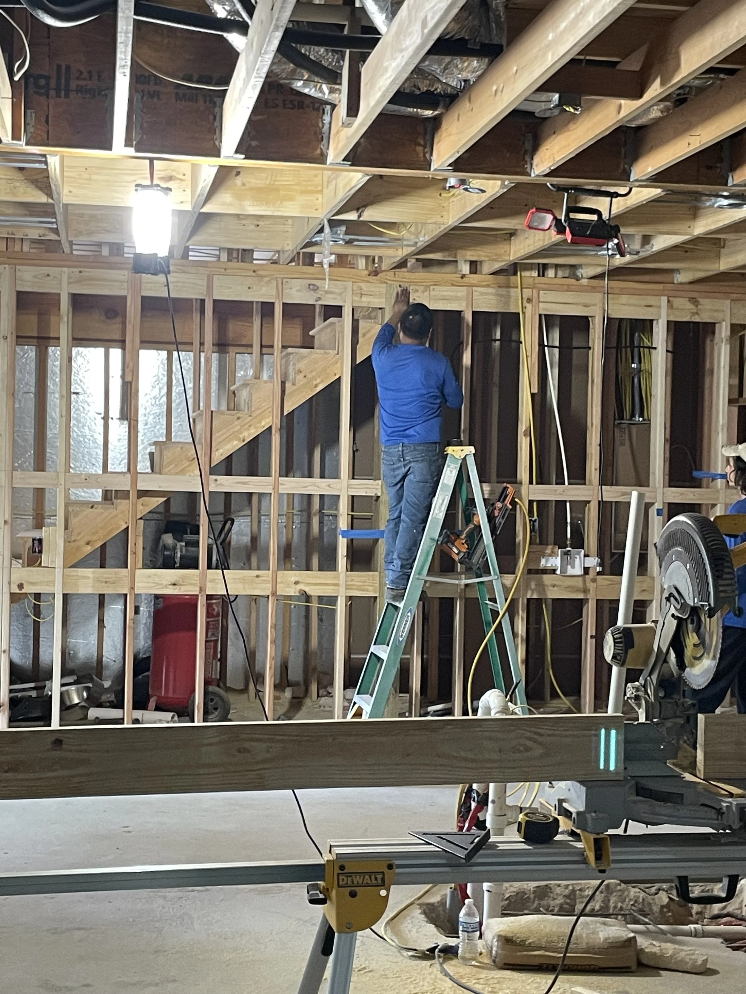 Construction workers are working inside a house under construction. One person is standing on a ladder, attaching or inspecting something on the ceiling. The room has exposed framing with wooden studs and ceiling beams, and construction tools and equ