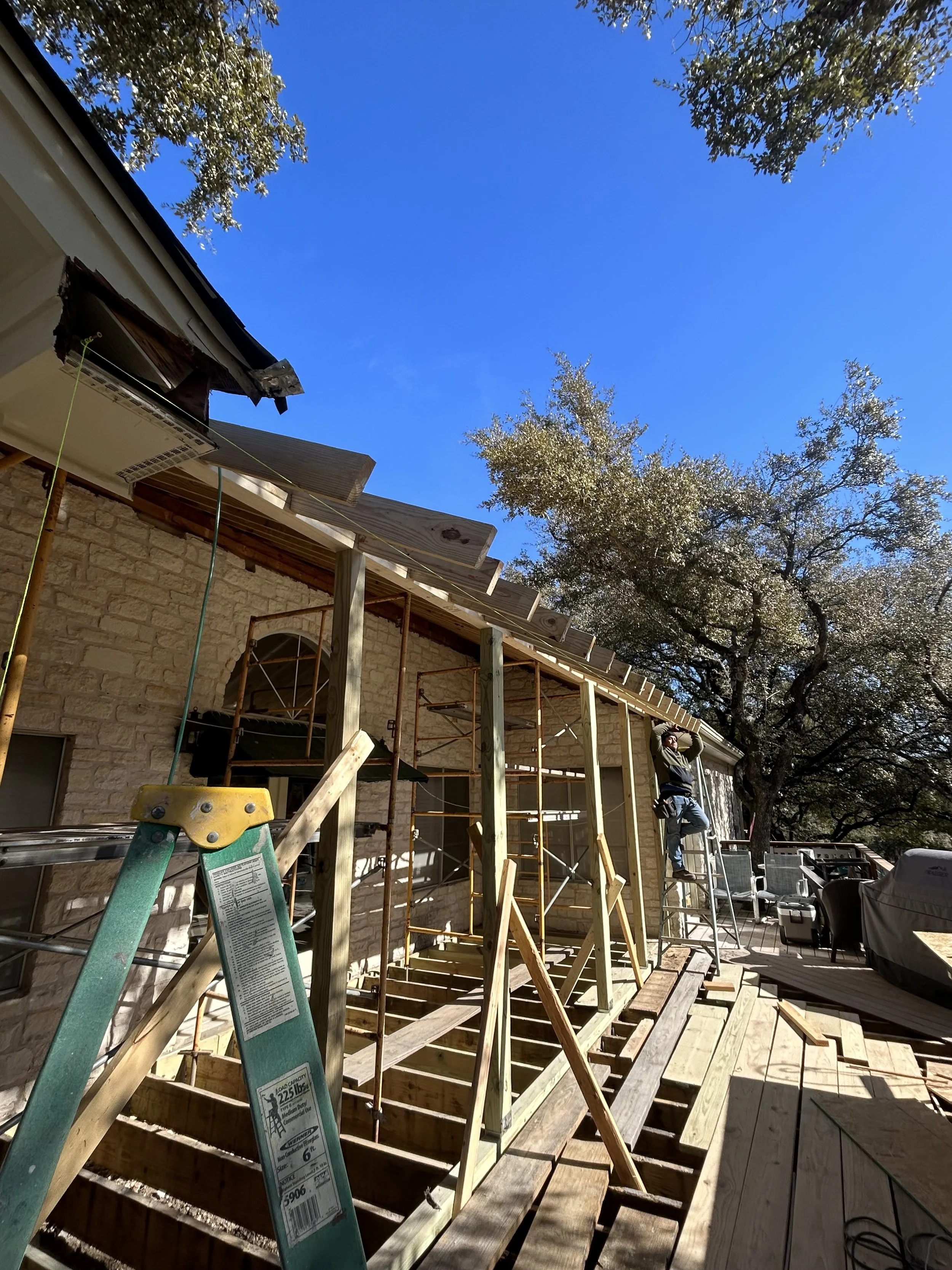 Construction worker using a ladder while working on a deck extension outside a house with brick siding, scaffolding, and wooden framing under a clear blue sky, surrounded by trees.