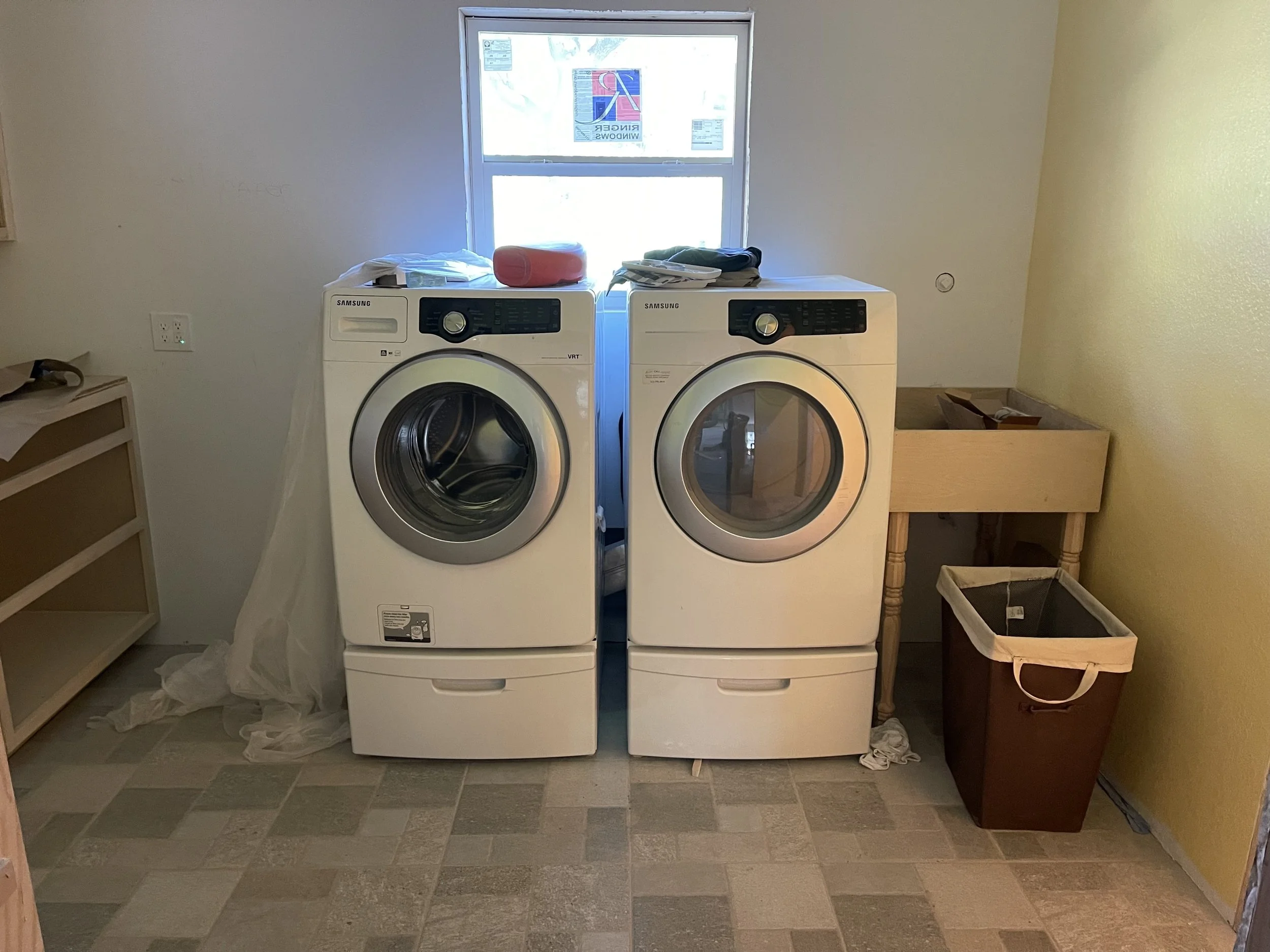 A laundry room with a window, two white Samsung front-loading washers side by side, and a wooden utility sink on the right. There is a laundry basket on the floor and a small wooden shelf to the left.