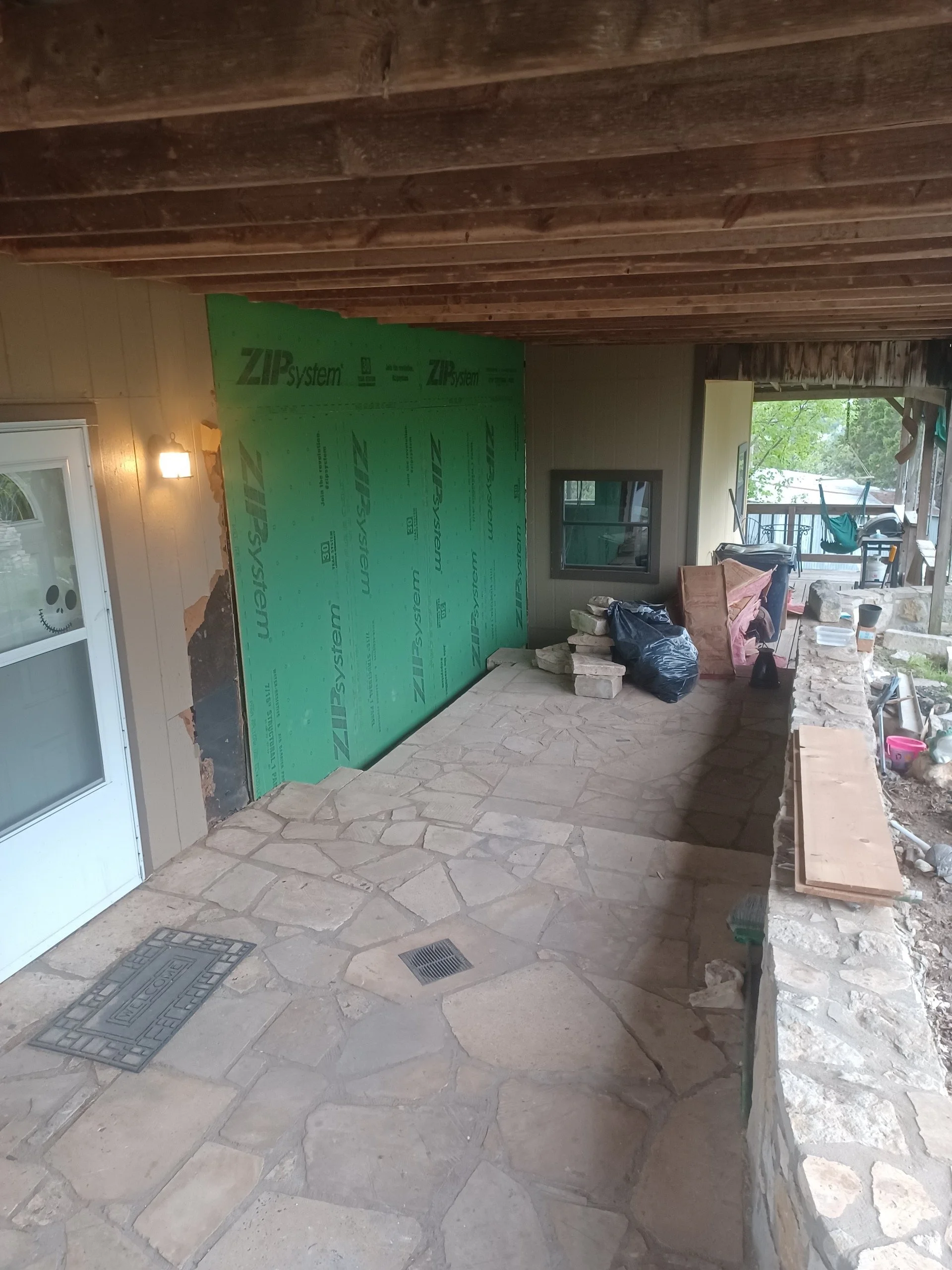 Back porch under renovation with stone flooring, unfinished wall with green insulation board, and construction materials scattered around.