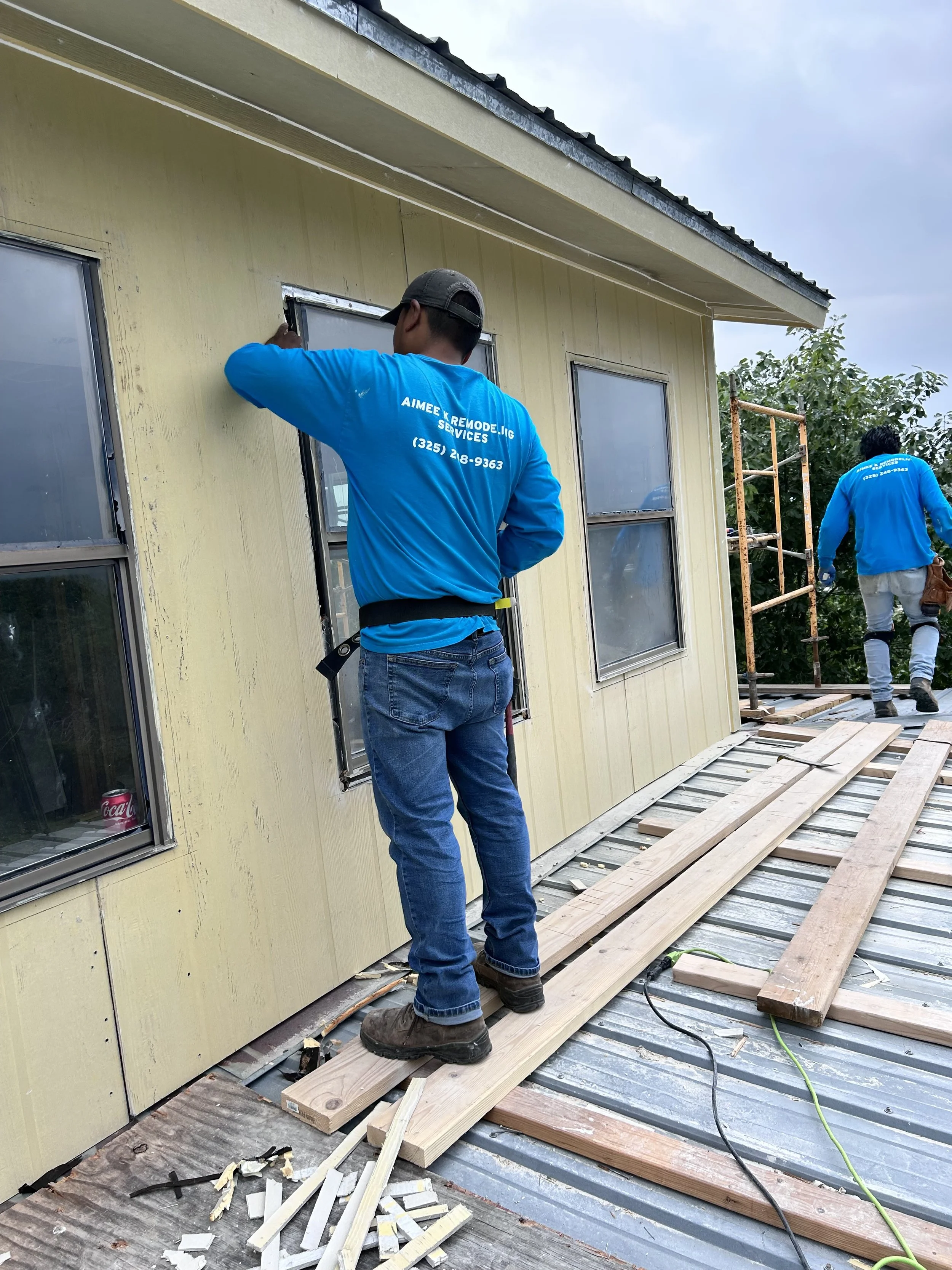 Workers installing windows on the exterior of a house with yellow siding during construction, with tools and wooden planks on the metal roof.