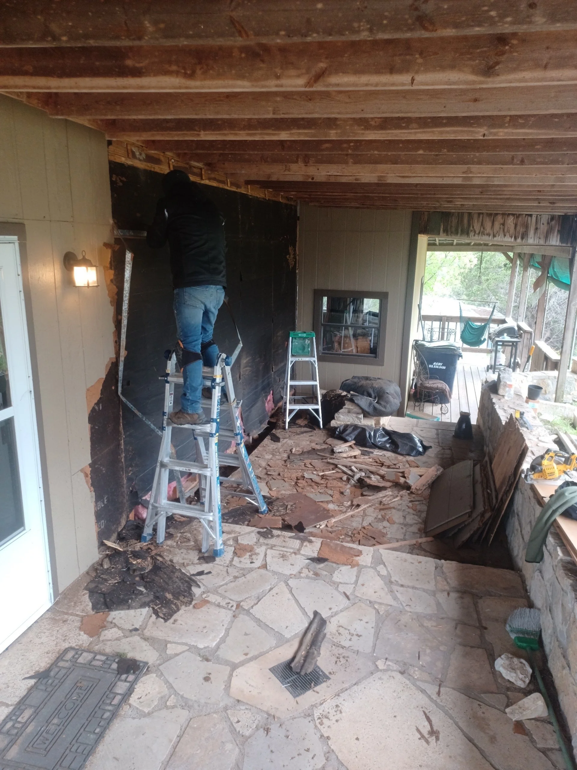 Person working on outdoor porch renovation, removing damaged flooring, with tools and construction materials scattered around.