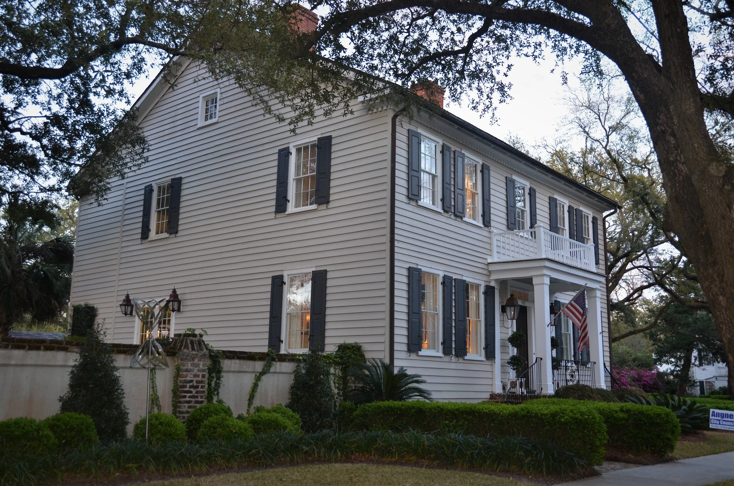 A white two-story house with black shutters, surrounded by trees and well-maintained bushes, with a small balcony and American flag near the front entrance.