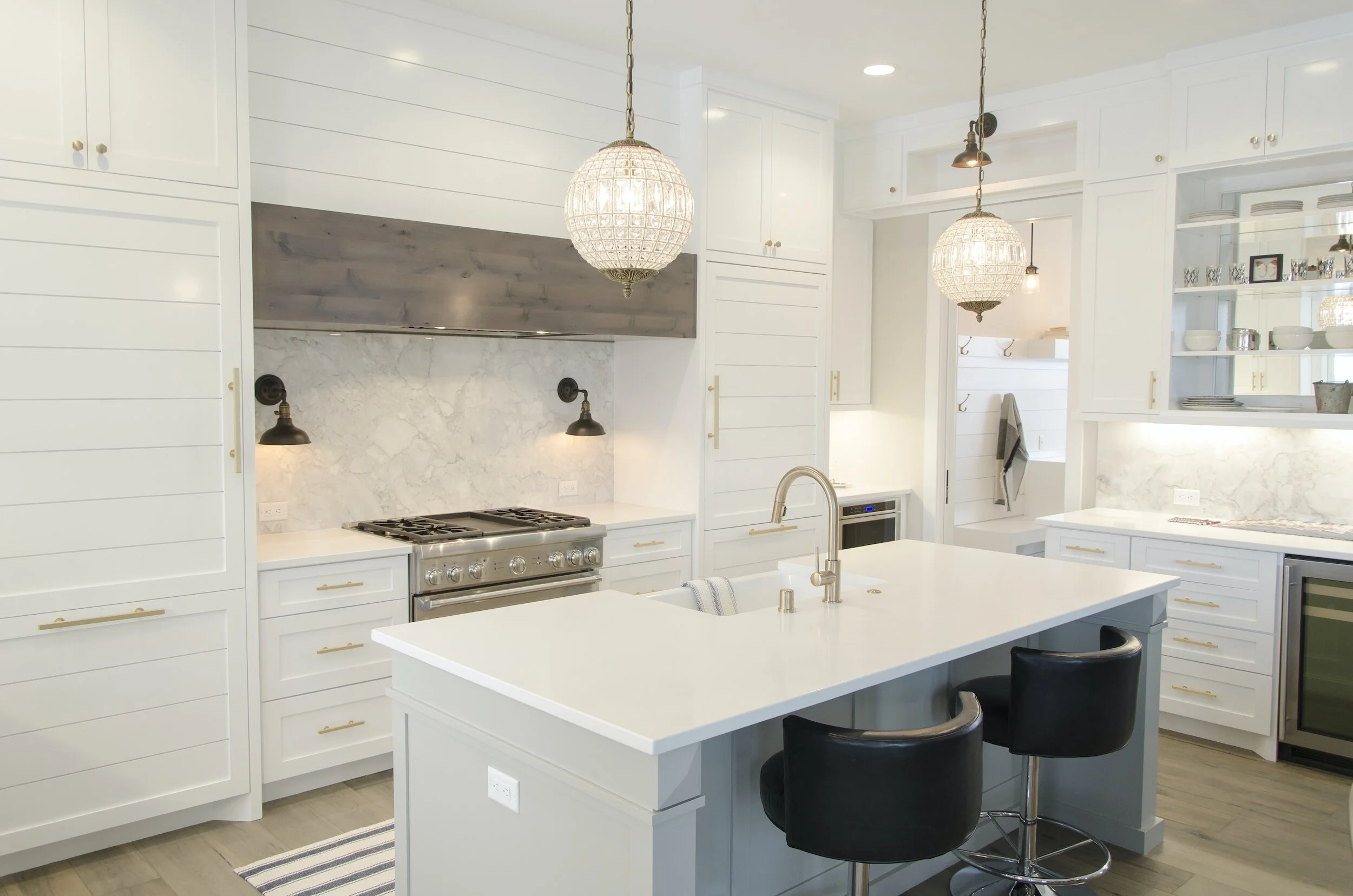 Modern white kitchen with a large central island, black bar stools, and pendant lighting, featuring sleek cabinetry, marble backsplash, and stainless steel appliances.