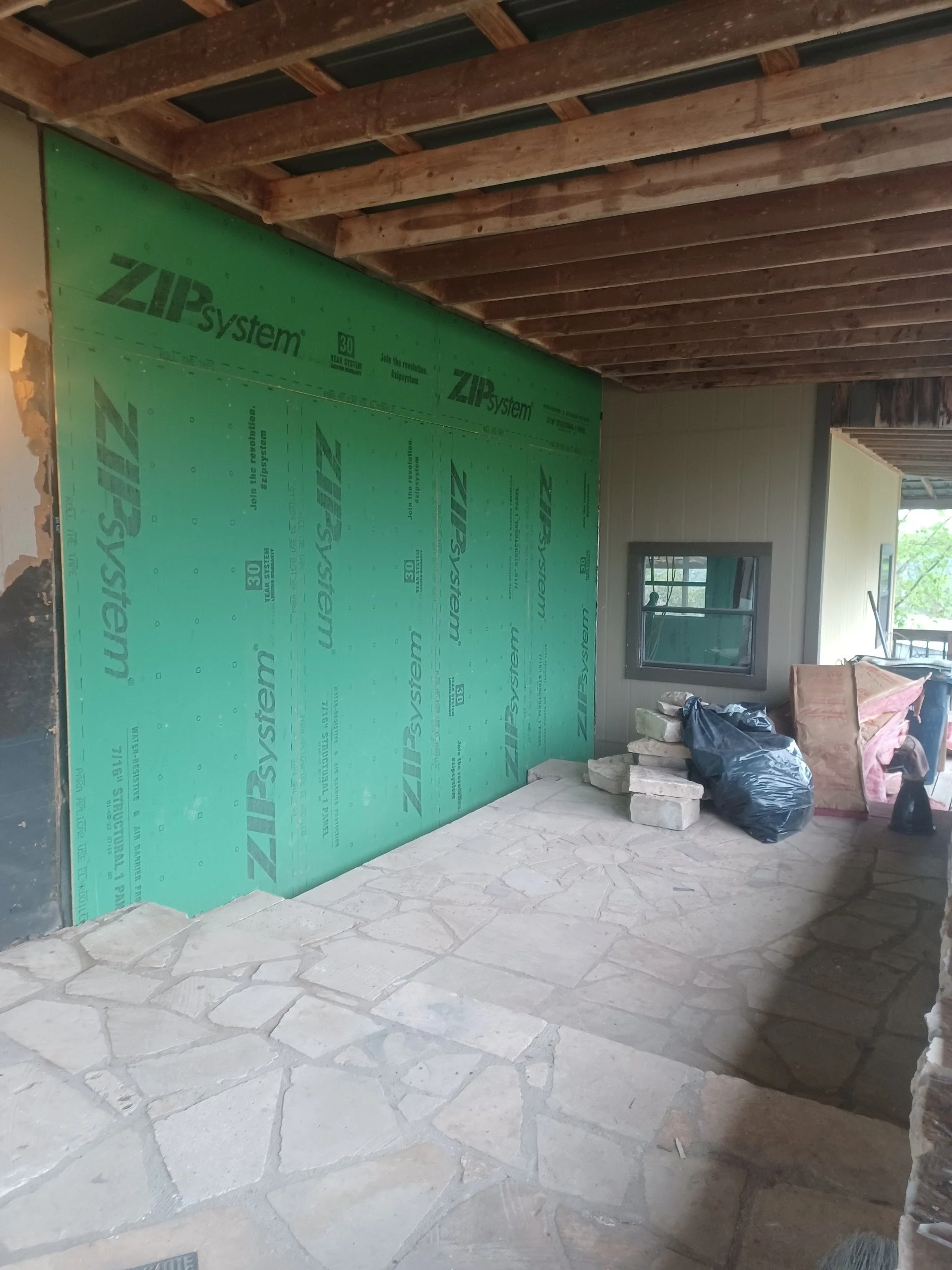 Construction site showing an interior wall with green ZIPSYSTEM sheathing, stone flooring, an unfinished wooden ceiling, a small window, and construction materials including bricks, a black trash bag, and other supplies.