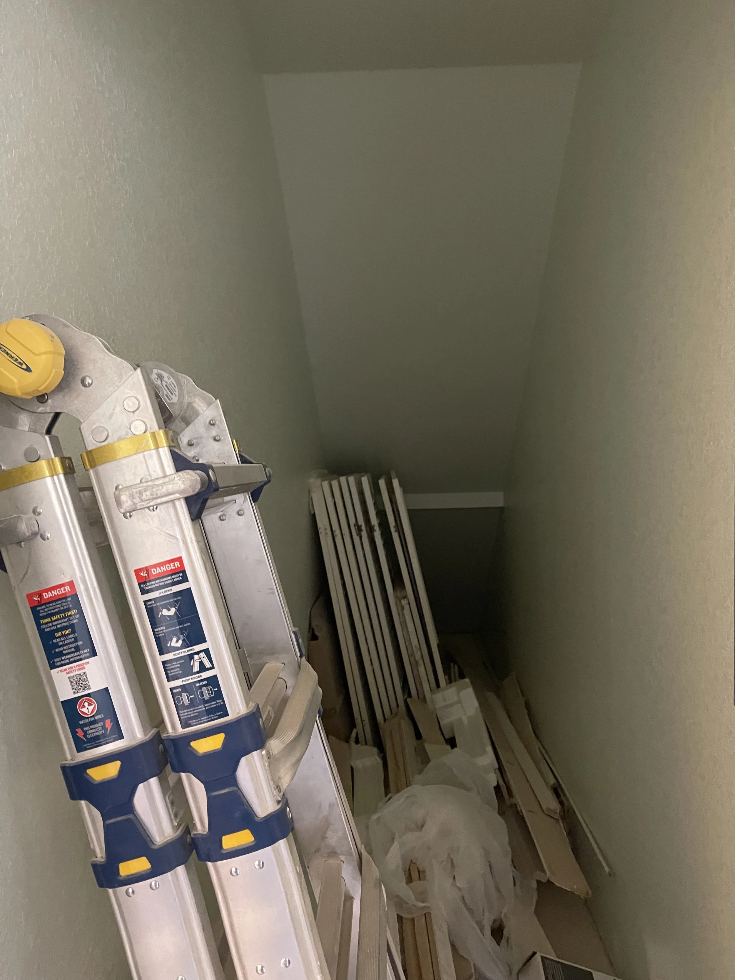 Attic with a ladder, white slats, cardboard, and plastic bags stored.