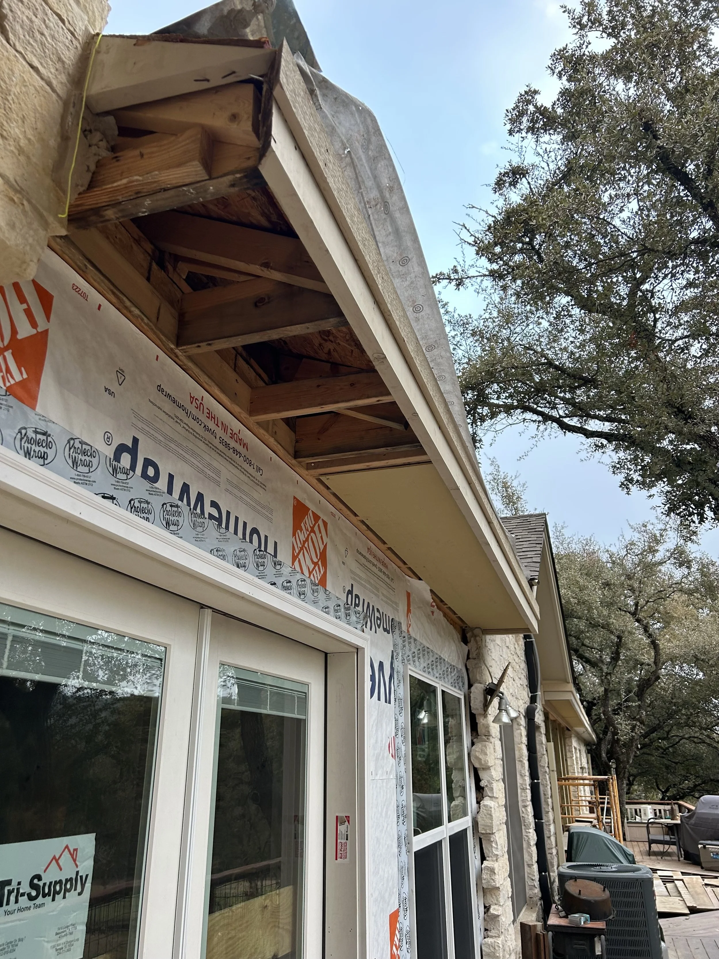 Side view of a house under construction with siding materials applied around windows, an overhang with exposed wooden framing, and trees in the background.