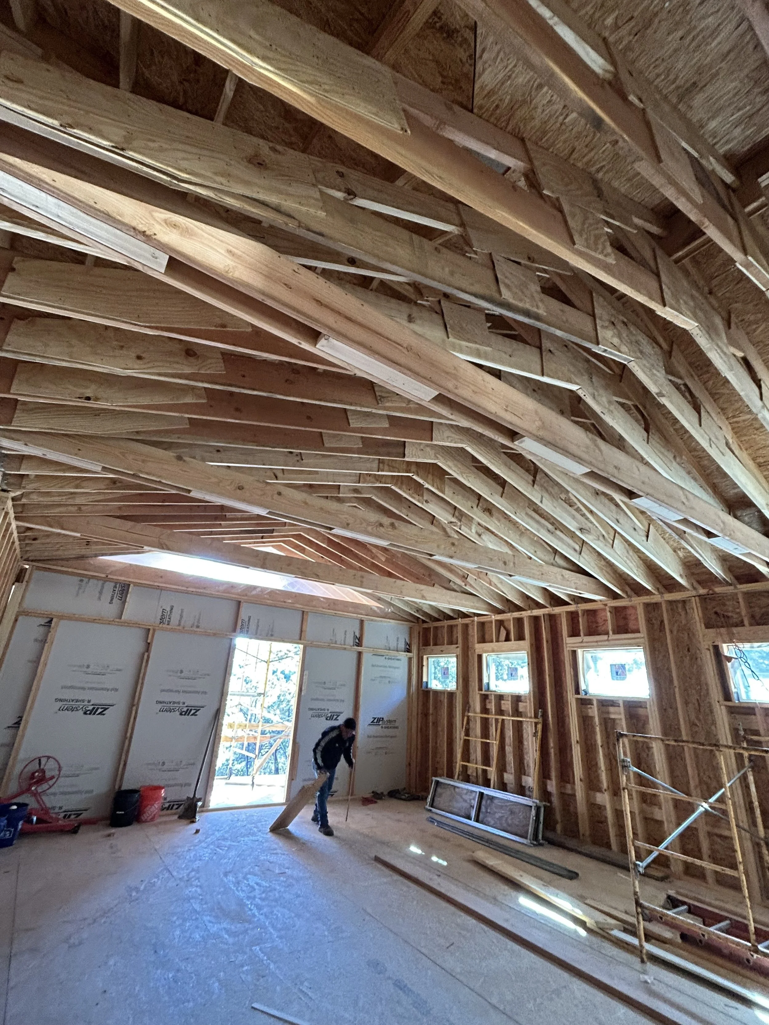 Interior of a house under construction with exposed wooden framing, a worker sweeping the floor, and construction tools and materials scattered around.