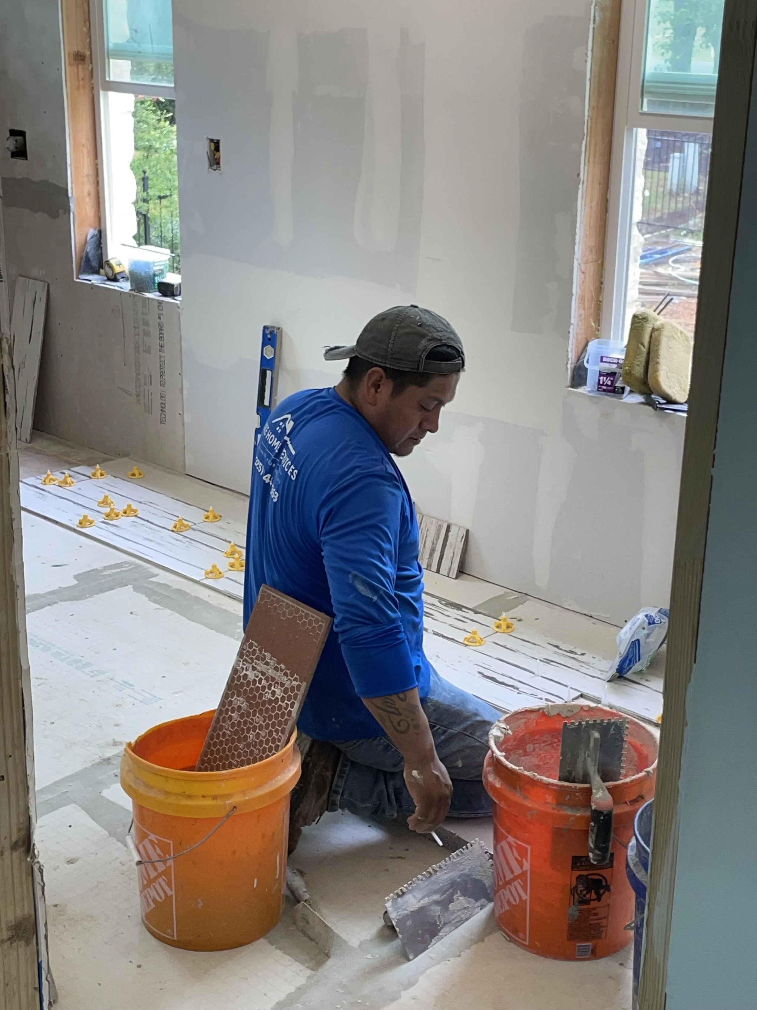 A man working on a home renovation, kneeling on the floor with buckets of tools and materials around him, with unfinished walls and windows in the background.
