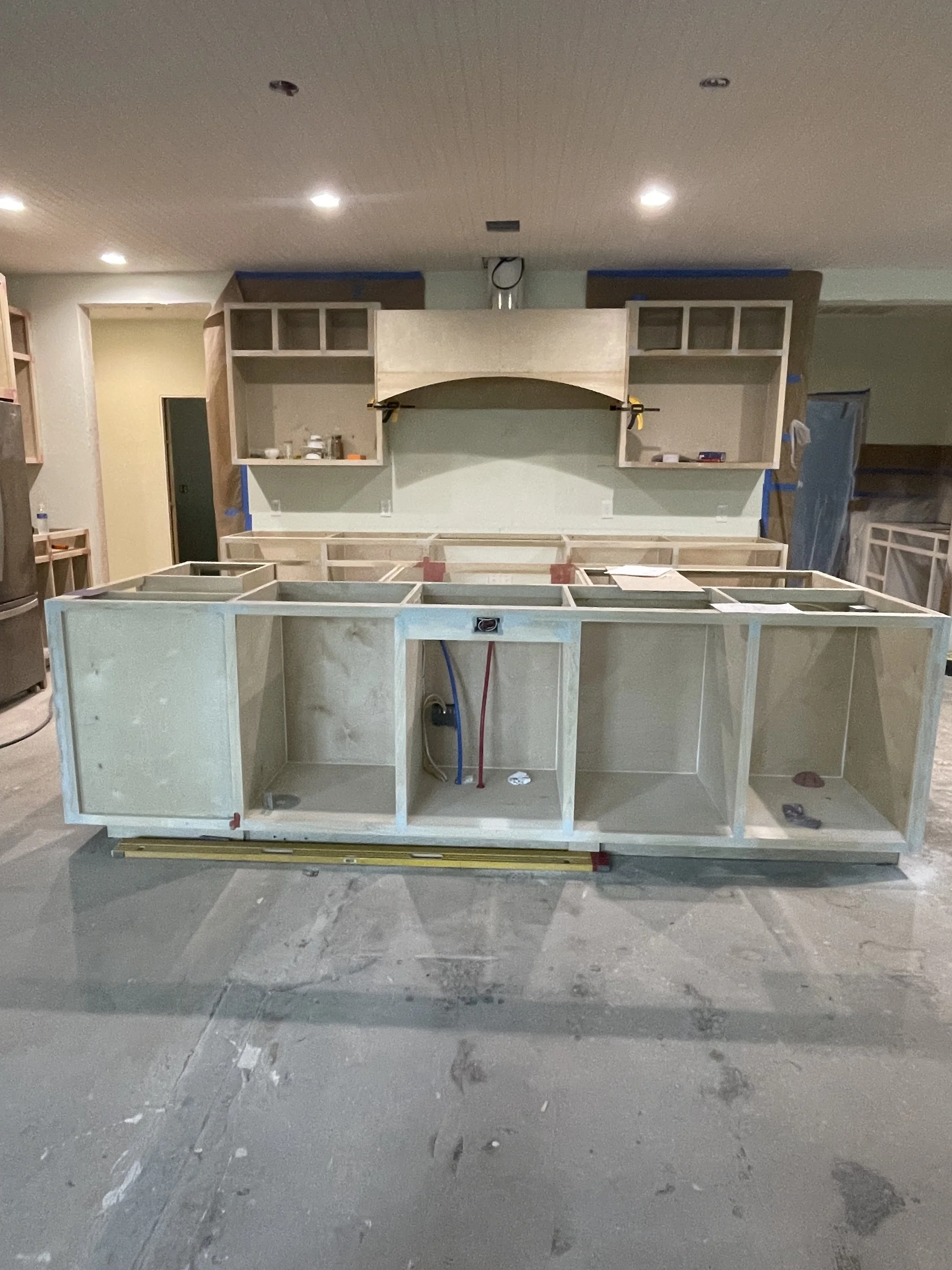 Unfinished kitchen cabinet installation with open wooden framing, plumbing pipes, and unfinished countertops in a room under construction.