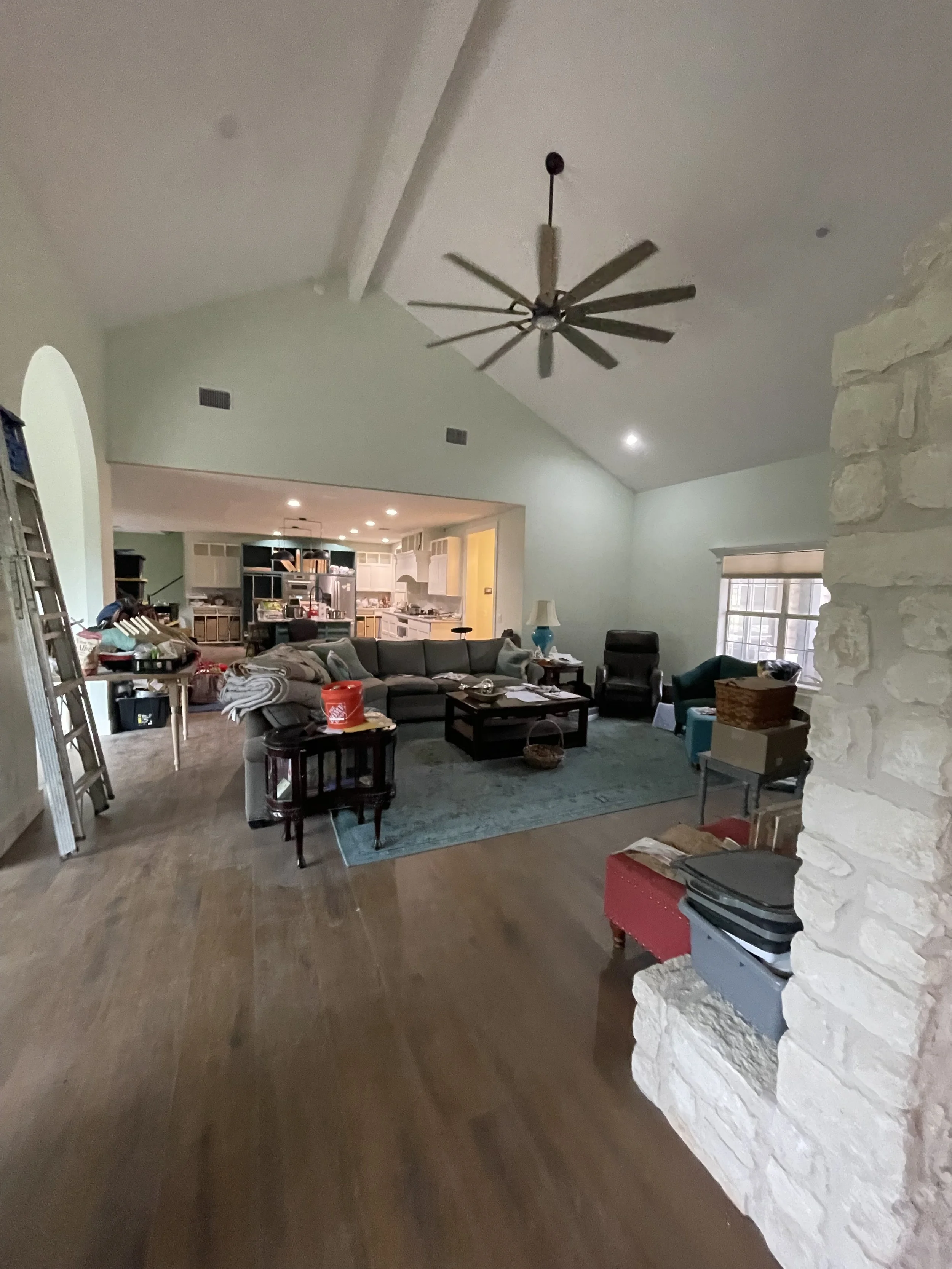 Living room with a vaulted ceiling, ceiling fan, sofa, armchair, coffee table, and a stone fireplace on the right. The room has hardwood floors, a window with blinds, and appears cluttered with various items.