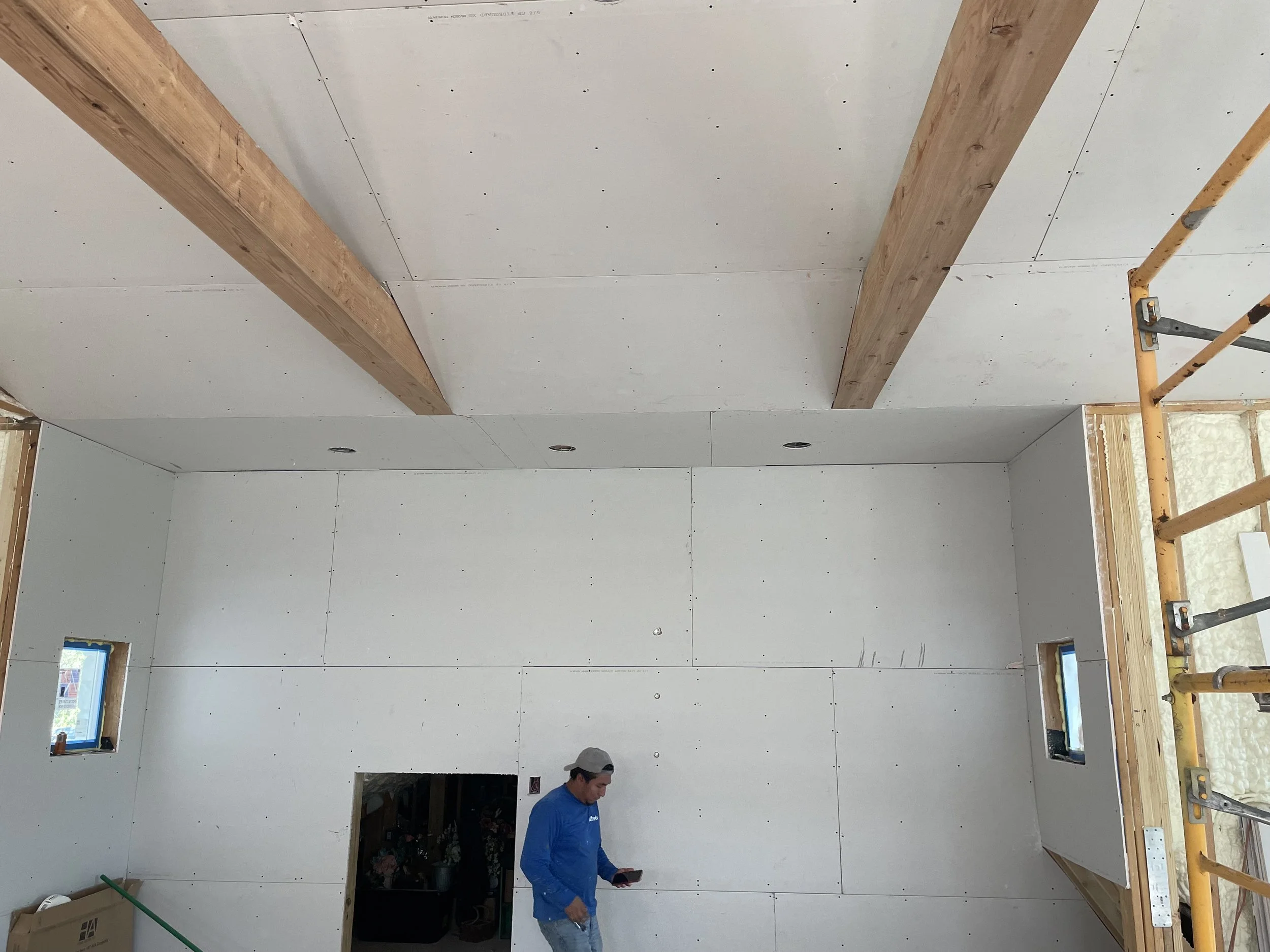 Interior of a building under construction with drywall walls, exposed wooden beams on the ceiling, a worker in a blue shirt and gray cap looking at his phone, and scaffolding on the right side.