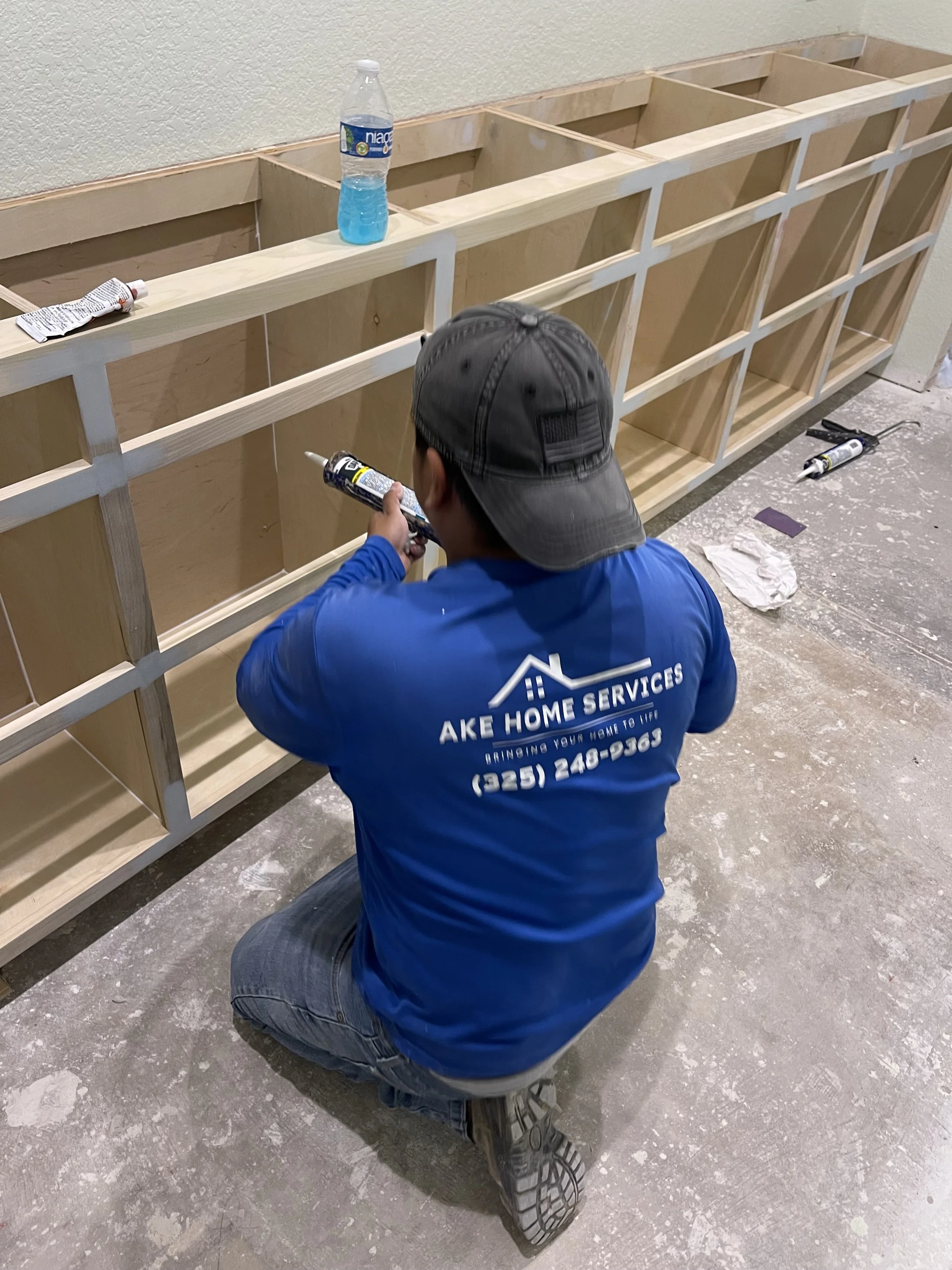 A worker in a blue shirt and black cap kneeling on the floor applying caulk to a wooden frame for a cabinet or shelf, with tools and materials scattered nearby.