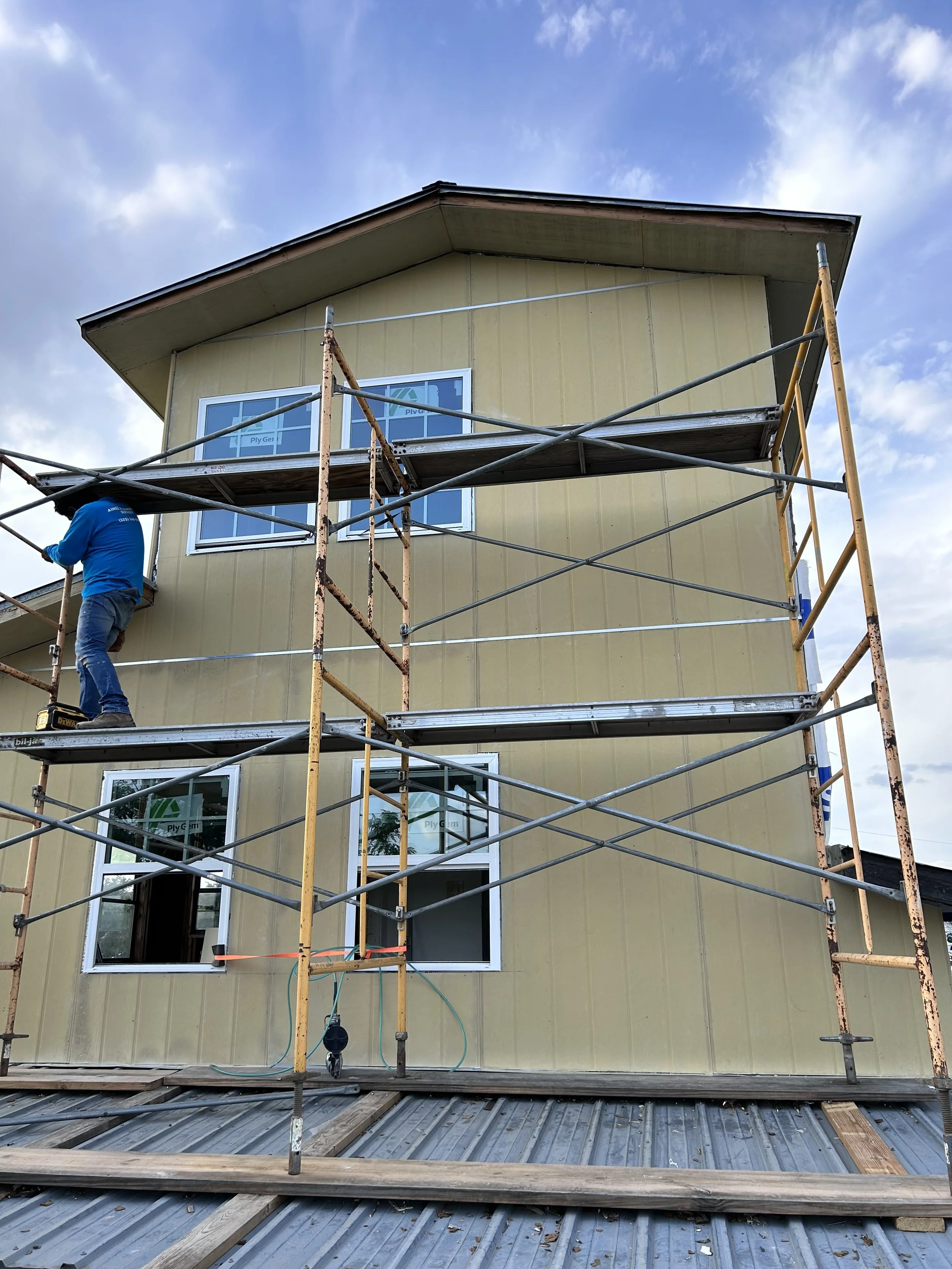 Construction worker on scaffolding working on a yellow house exterior with multiple windows, under a partly cloudy sky.