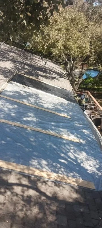 A person working on a roof with a tarp covering the surface, surrounded by trees and a backyard with a swimming pool in the background.