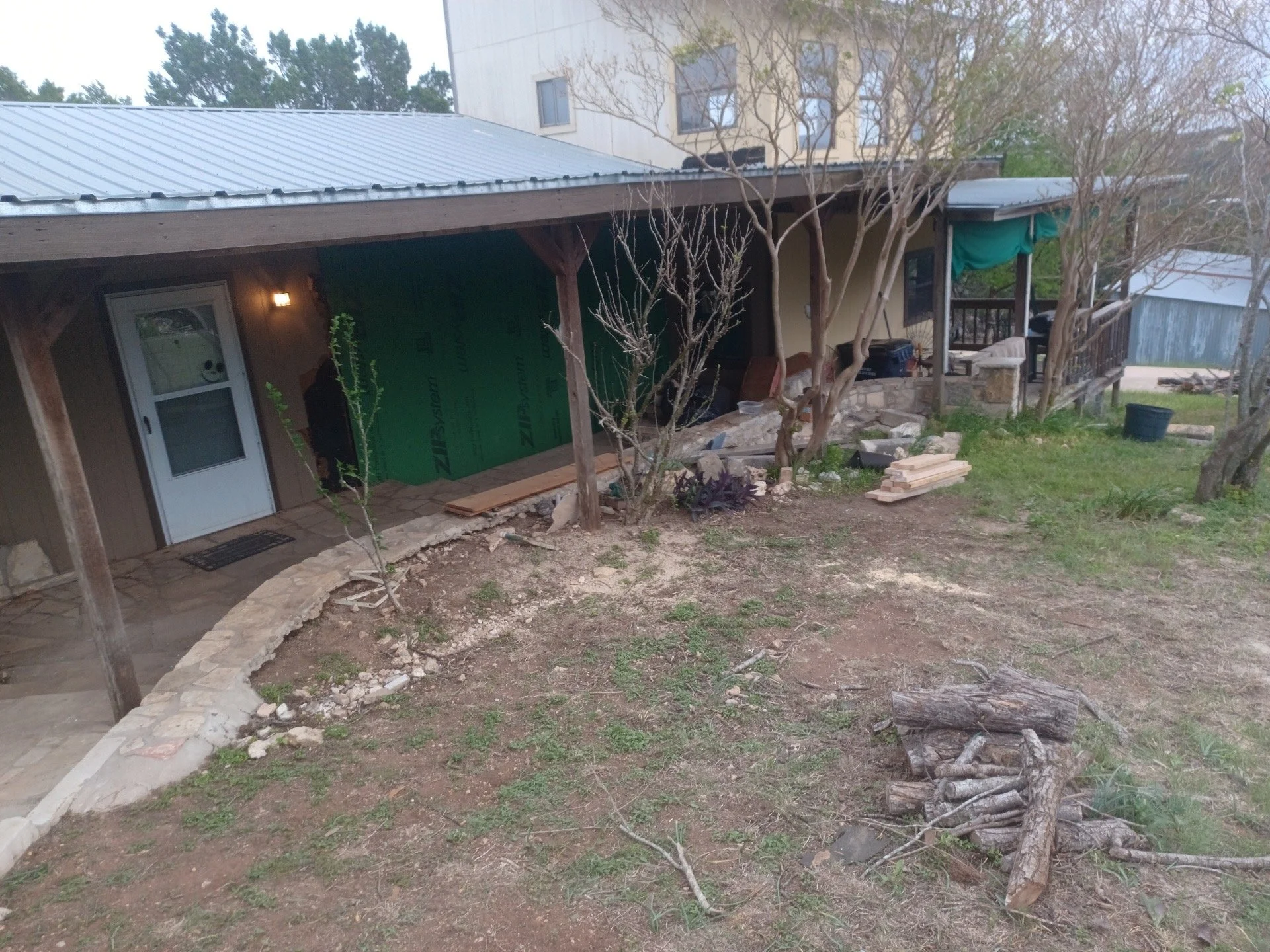 Backyard of a house under construction, with a covered porch, small trees, a pile of chopped wood, and construction materials scattered on the ground.
