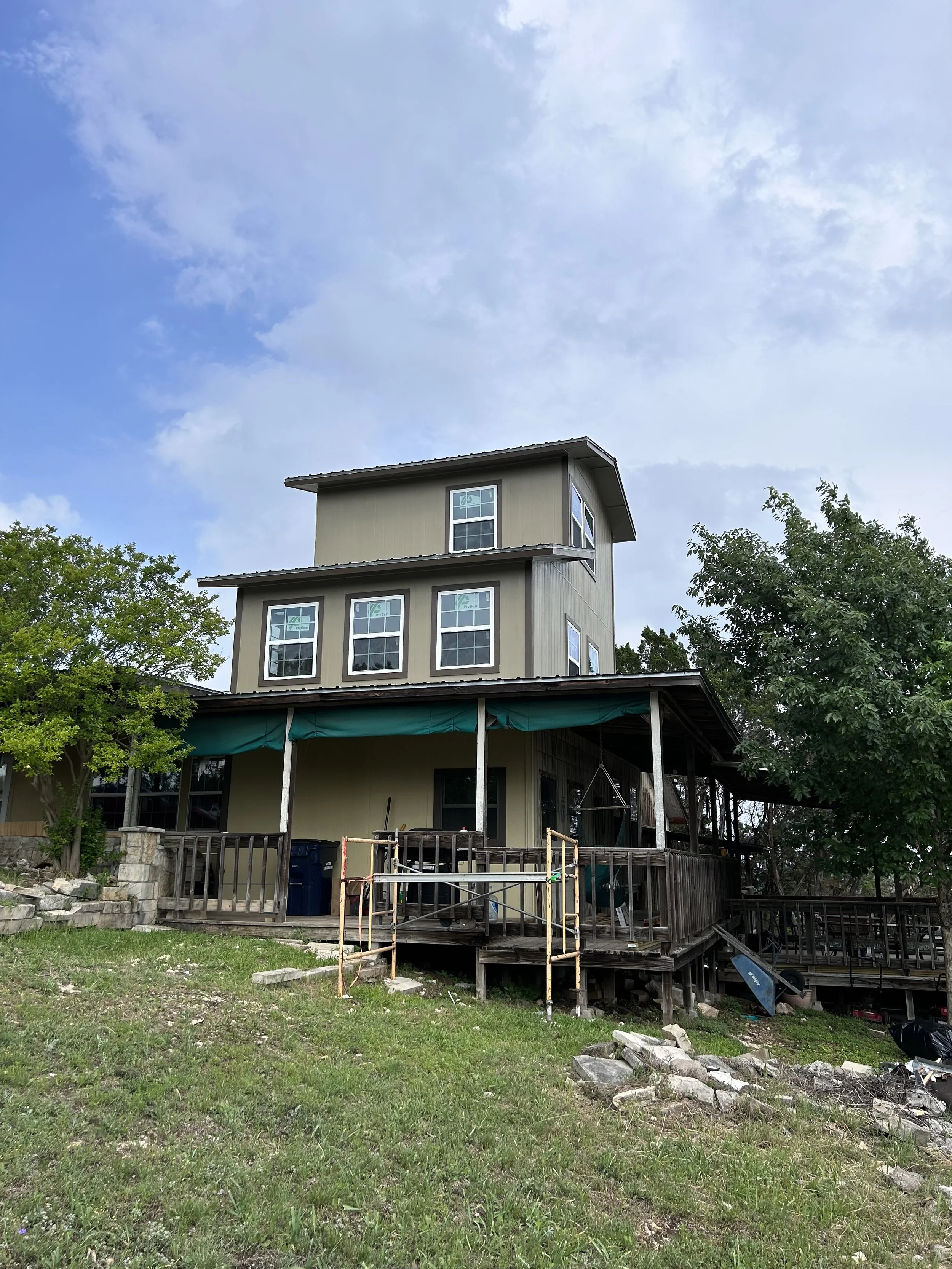 A house with an unfinished wooden porch, scaffolding, scaffolding, and surrounded by trees and grass, under a partly cloudy sky.