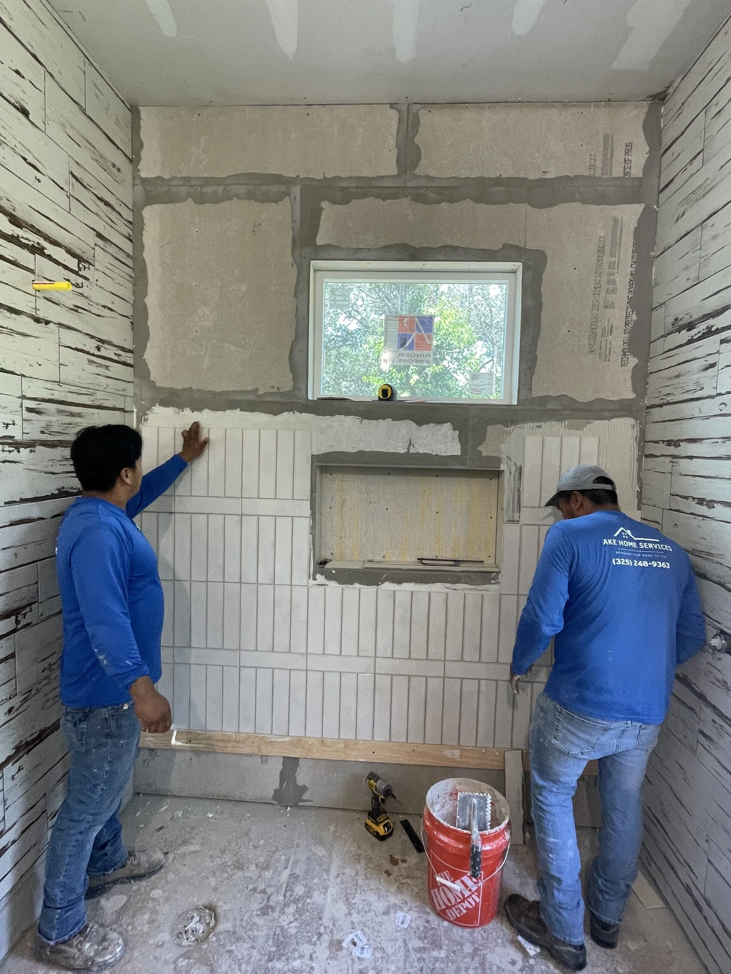 Two workers installing white tiled wall in a room under construction, with a small window above.