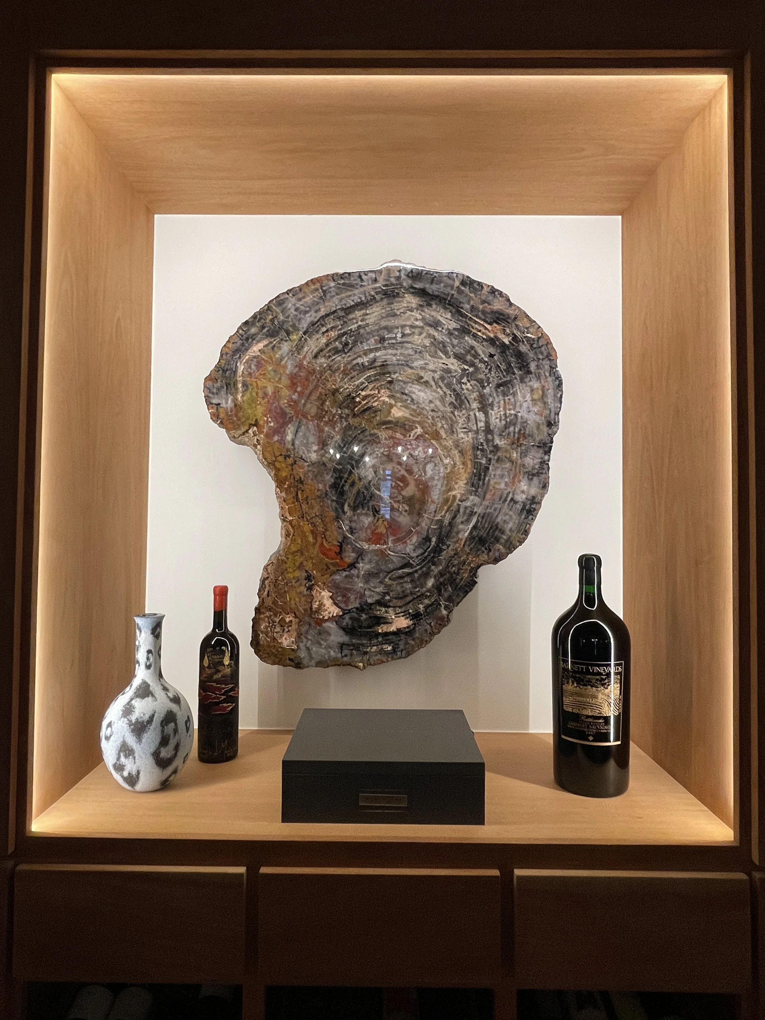 Illuminated wooden display shelf with a large fossilized ammonite, two wine bottles, and a decorative ceramic vase