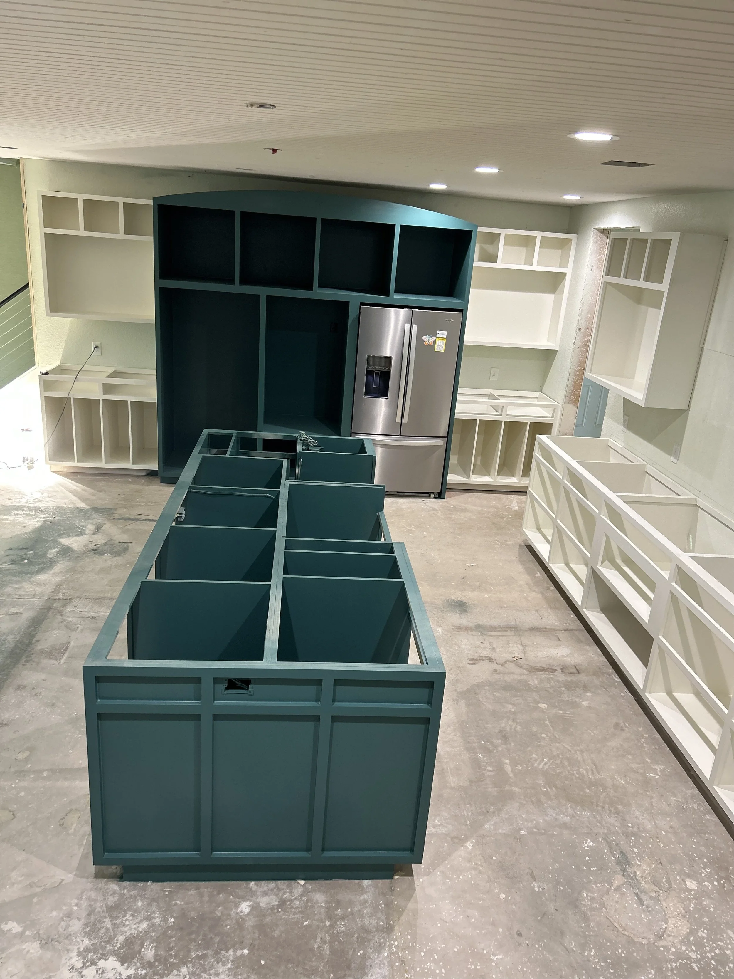 Interior of a kitchen under construction with teal and white cabinets and a stainless steel refrigerator.