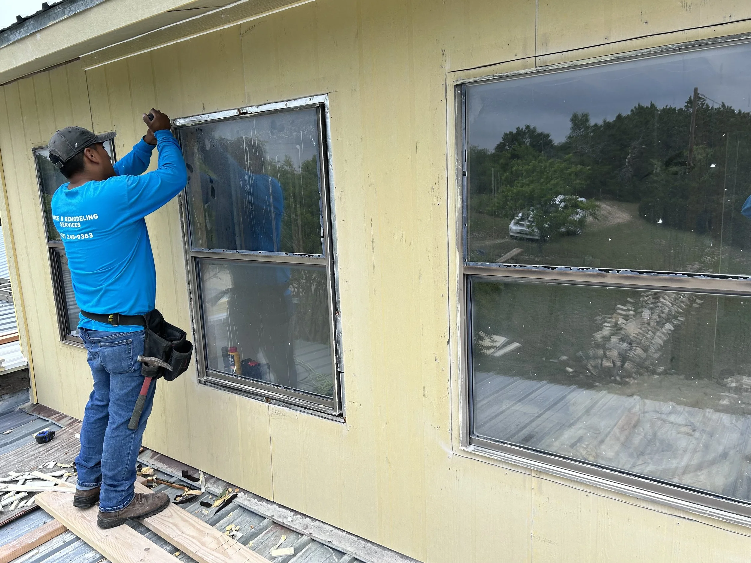 A worker installing or repairing a window on the exterior wall of a building. The worker is wearing a blue shirt, jeans, and a cap, and is standing on a wooden platform. The building's exterior is yellow, and there are two large windows visible.