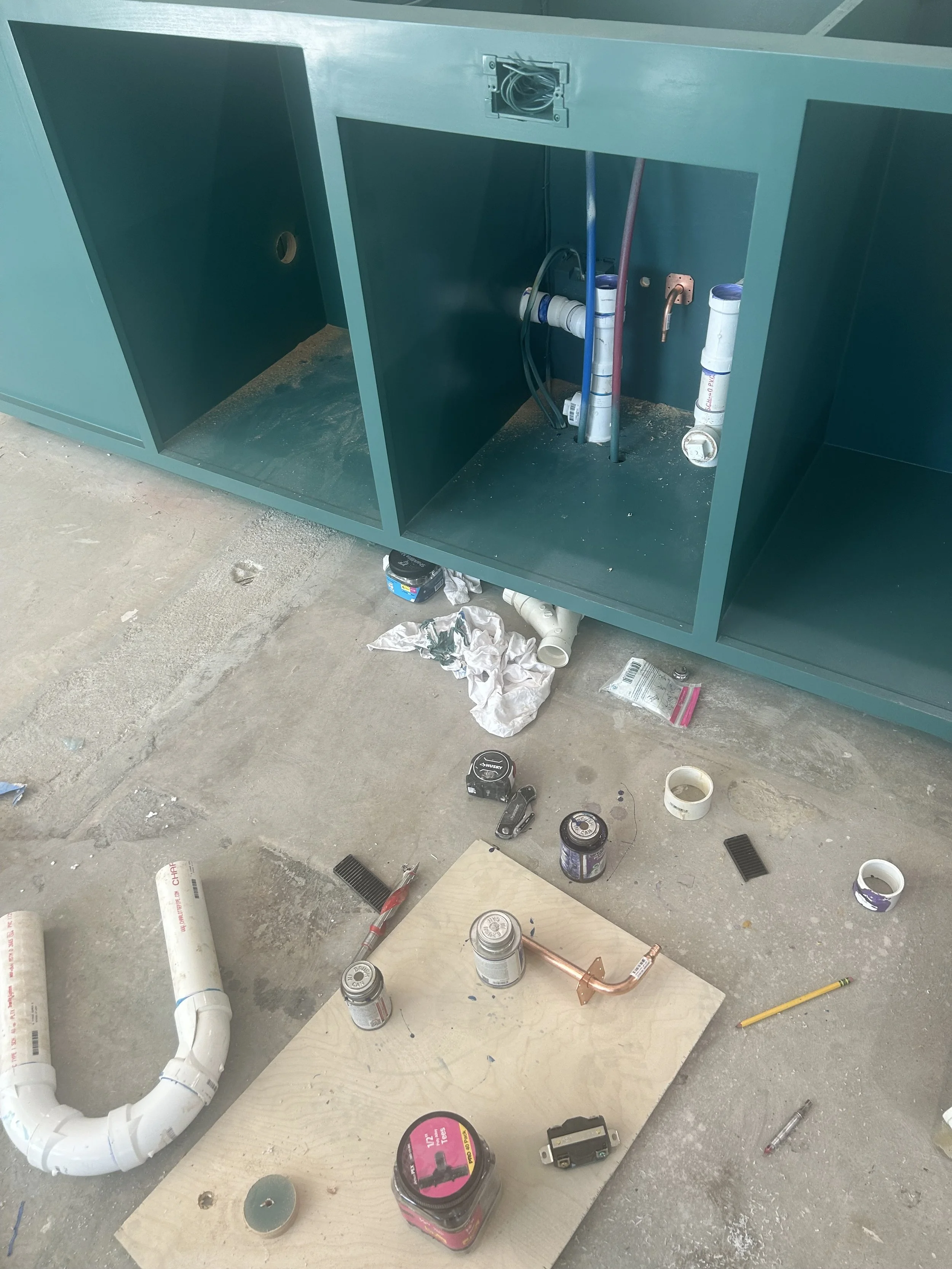 Under construction or repair scene showing plumbing pipes inside a green cabinet with tools, paint cans, pipes, a pipe cutter, and other supplies on the floor.