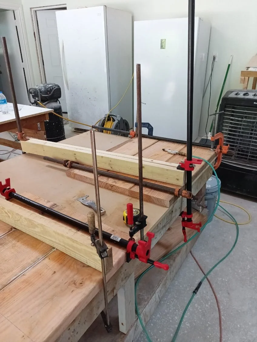 A woodworking or DIY project in progress with wood pieces held together by clamps on a workbench, in a garage or workshop setting. Tools, a measuring tape, and a corded air compressor are visible.