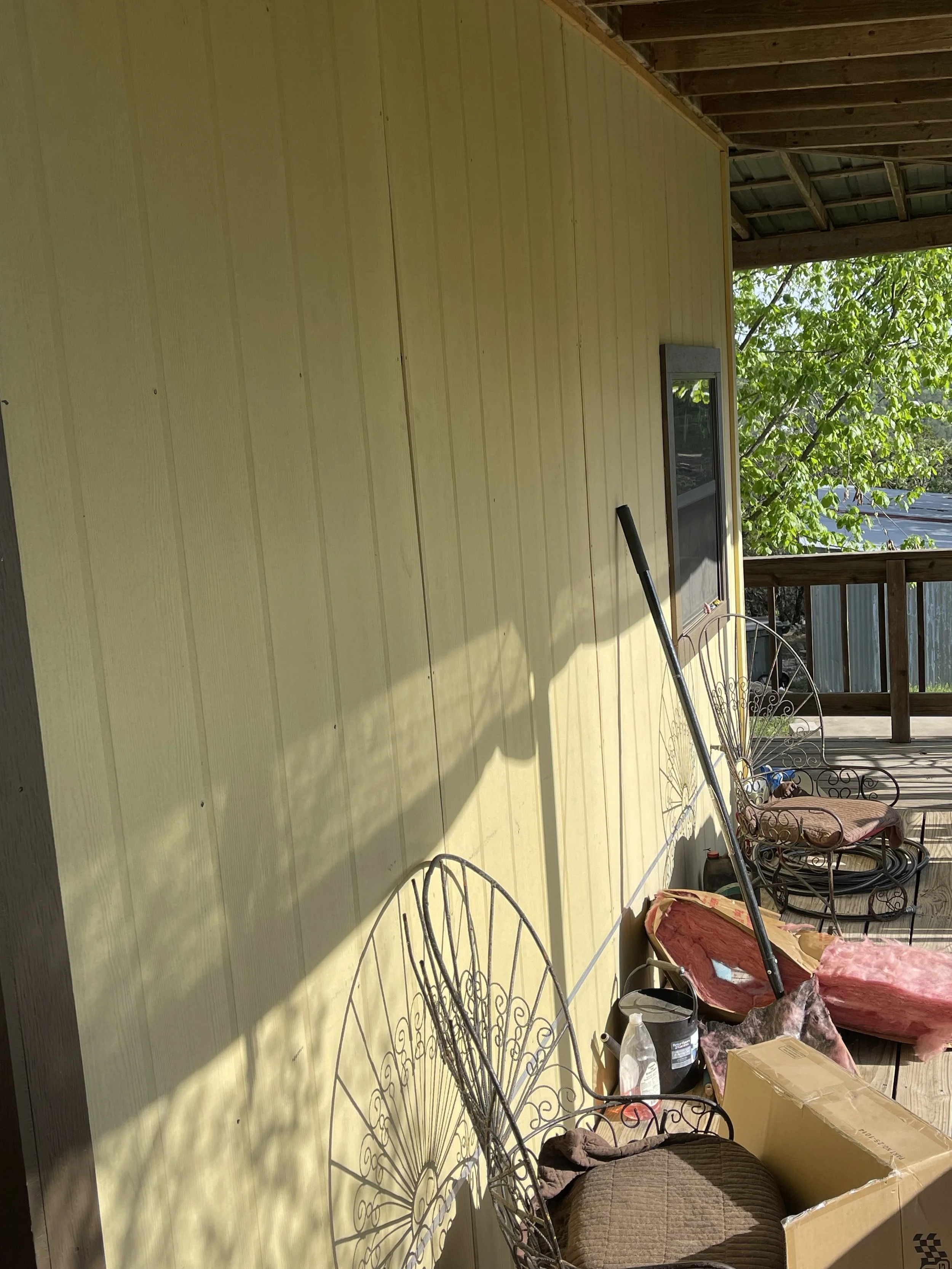 A covered porch area with yellow siding, metal chairs, cushions, a cardboard box, and a patio rake. Sunlight and shadows are visible.