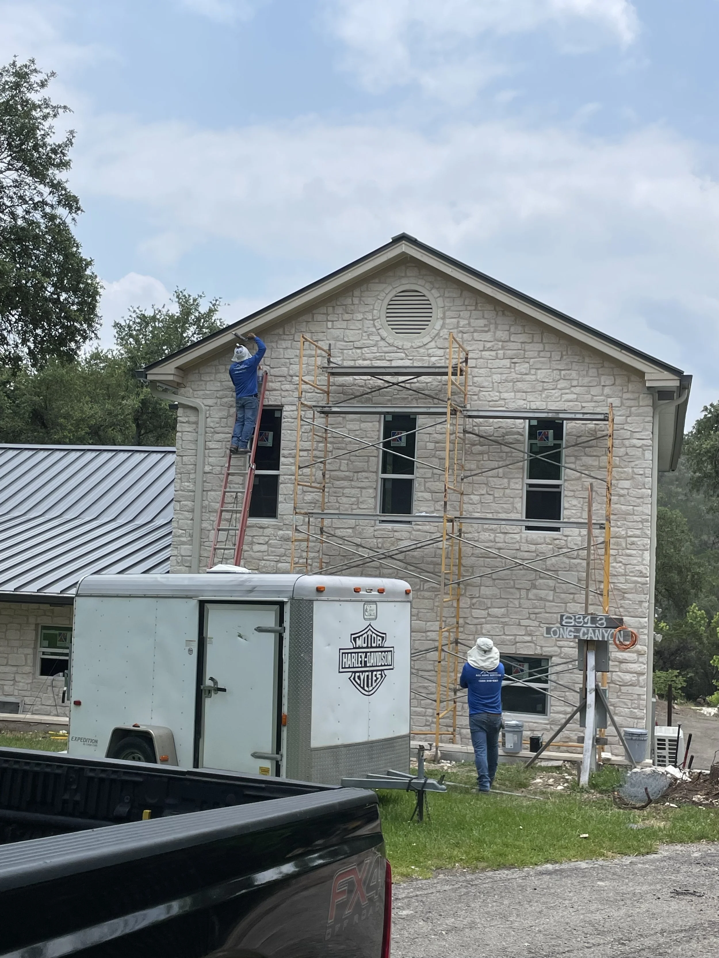 Two workers are painting the upper exterior wall of a two-story house made of stone. One worker is standing on a ladder, and the other is on the ground operating a spray gun. There is scaffolding on the house, a trailer with a Harley-Davidson logo, a