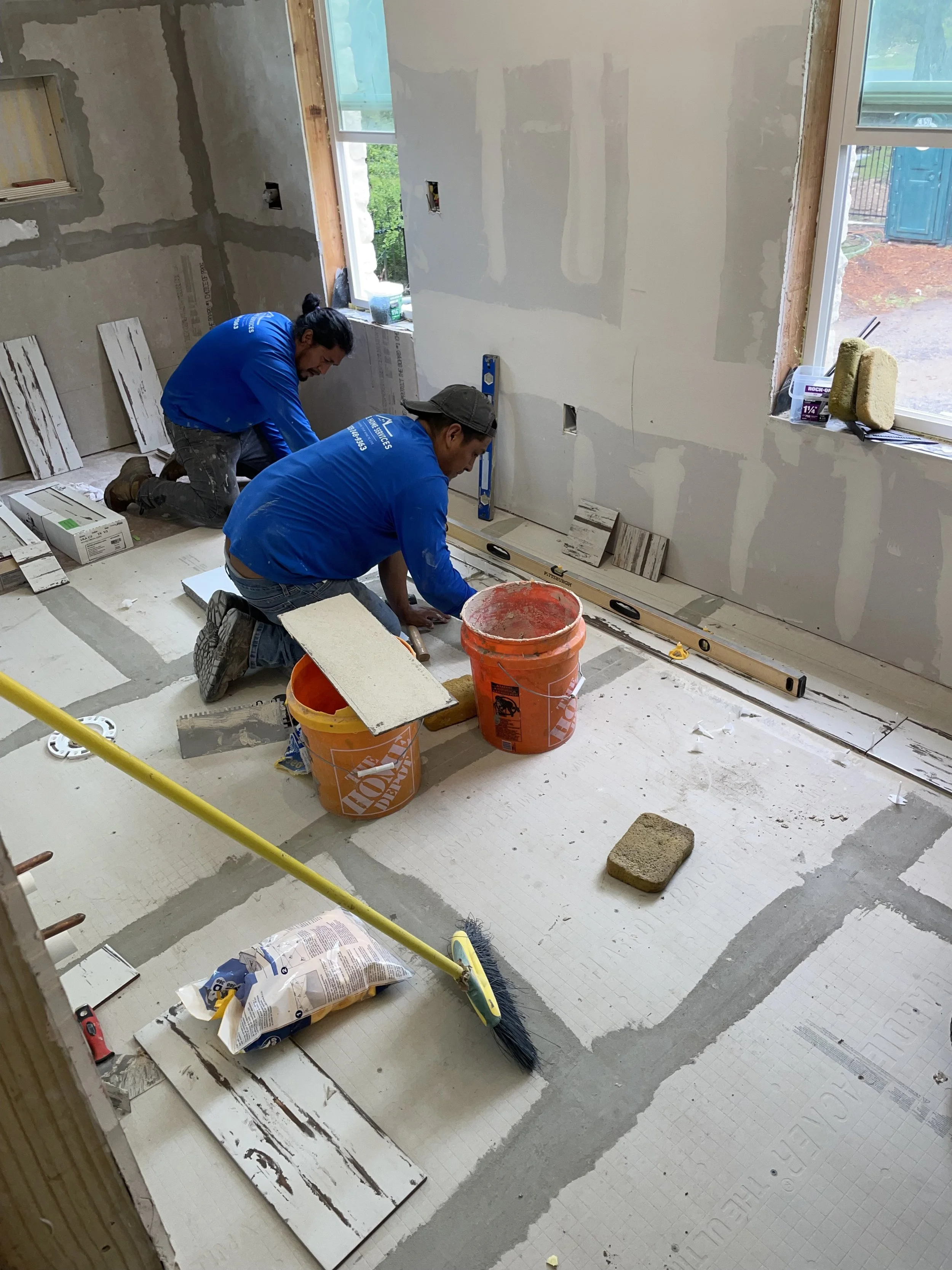 Two workers installing tile flooring in a room under construction, with tools and supplies around them.