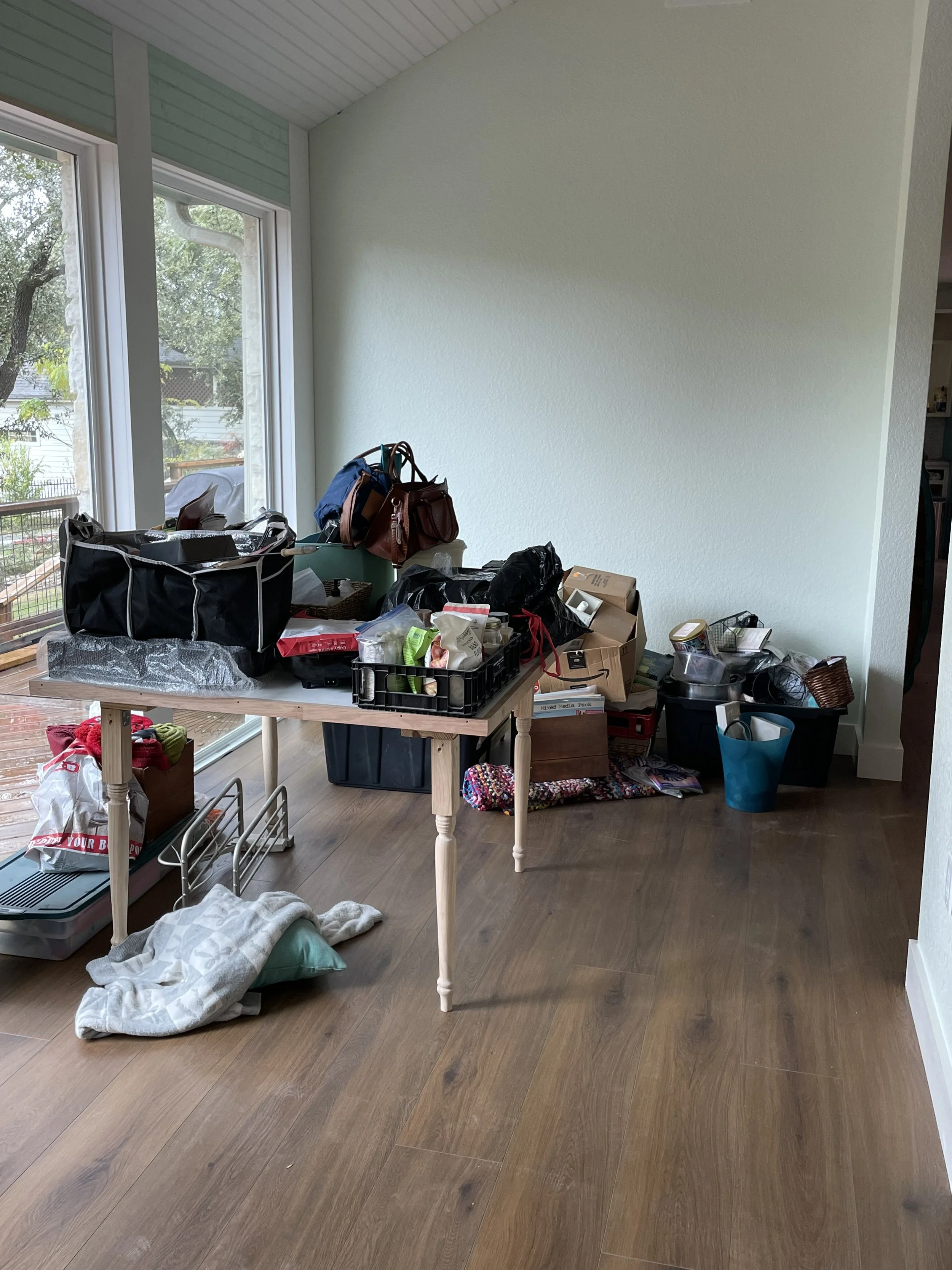Room with a table covered in various bags, boxes, and clutter; near large windows with a view of trees outside.