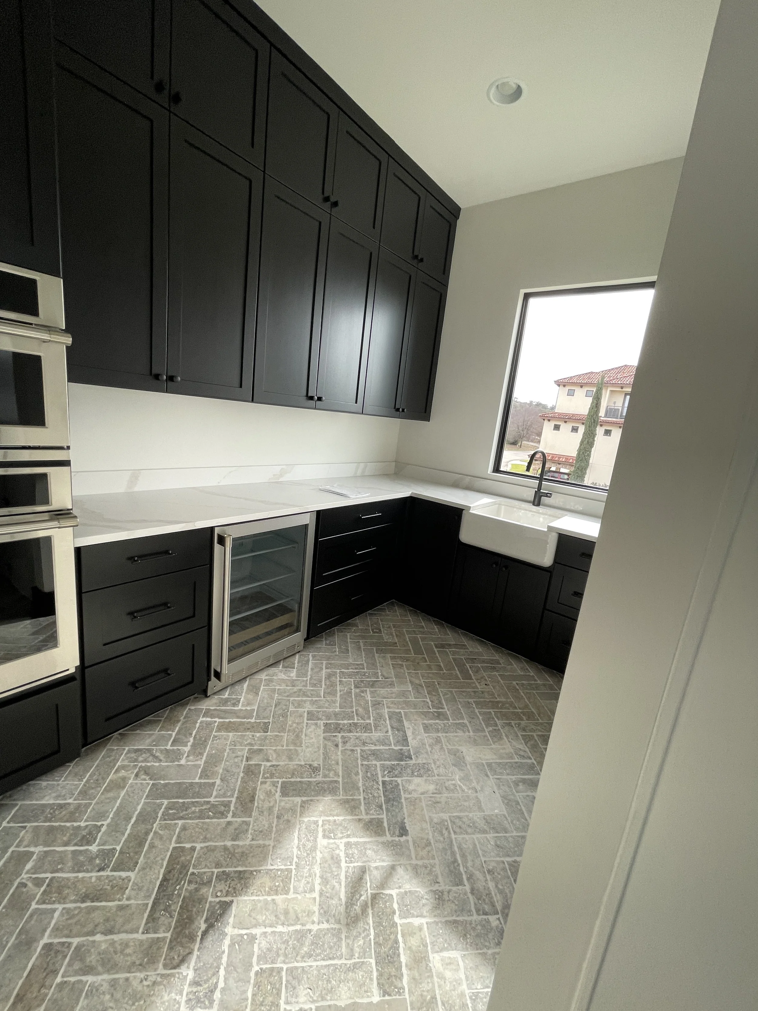 Modern kitchen with black cabinets, white countertops, farmhouse sink, stainless steel appliances, and gray stone tile flooring.