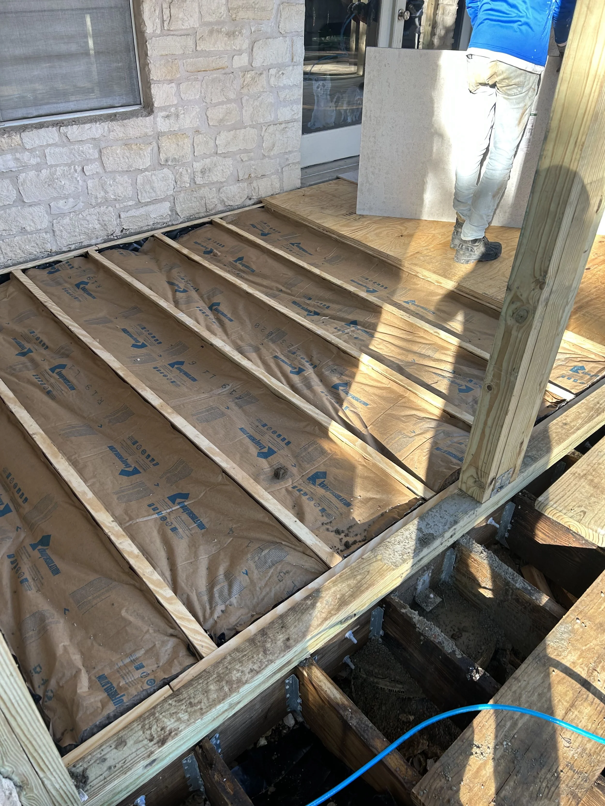 Construction worker installing insulation and plywood on a wood deck frame outside a building with a stone exterior wall.