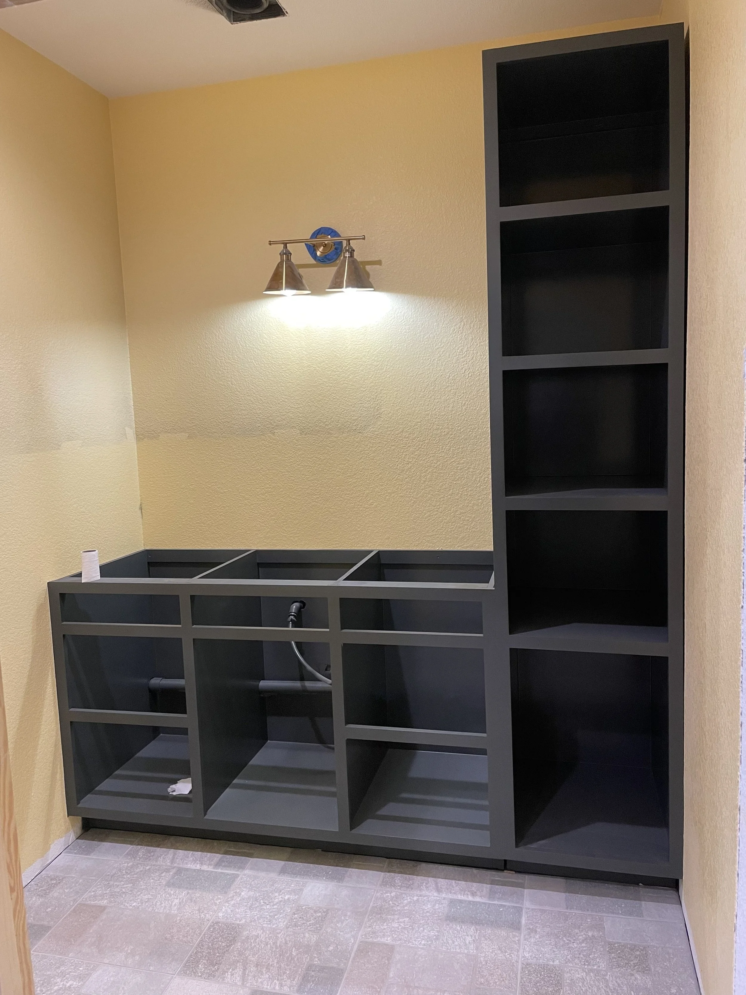 Black kitchen cabinet with open shelves next to a beige wall with a light fixture, in an unfinished room.