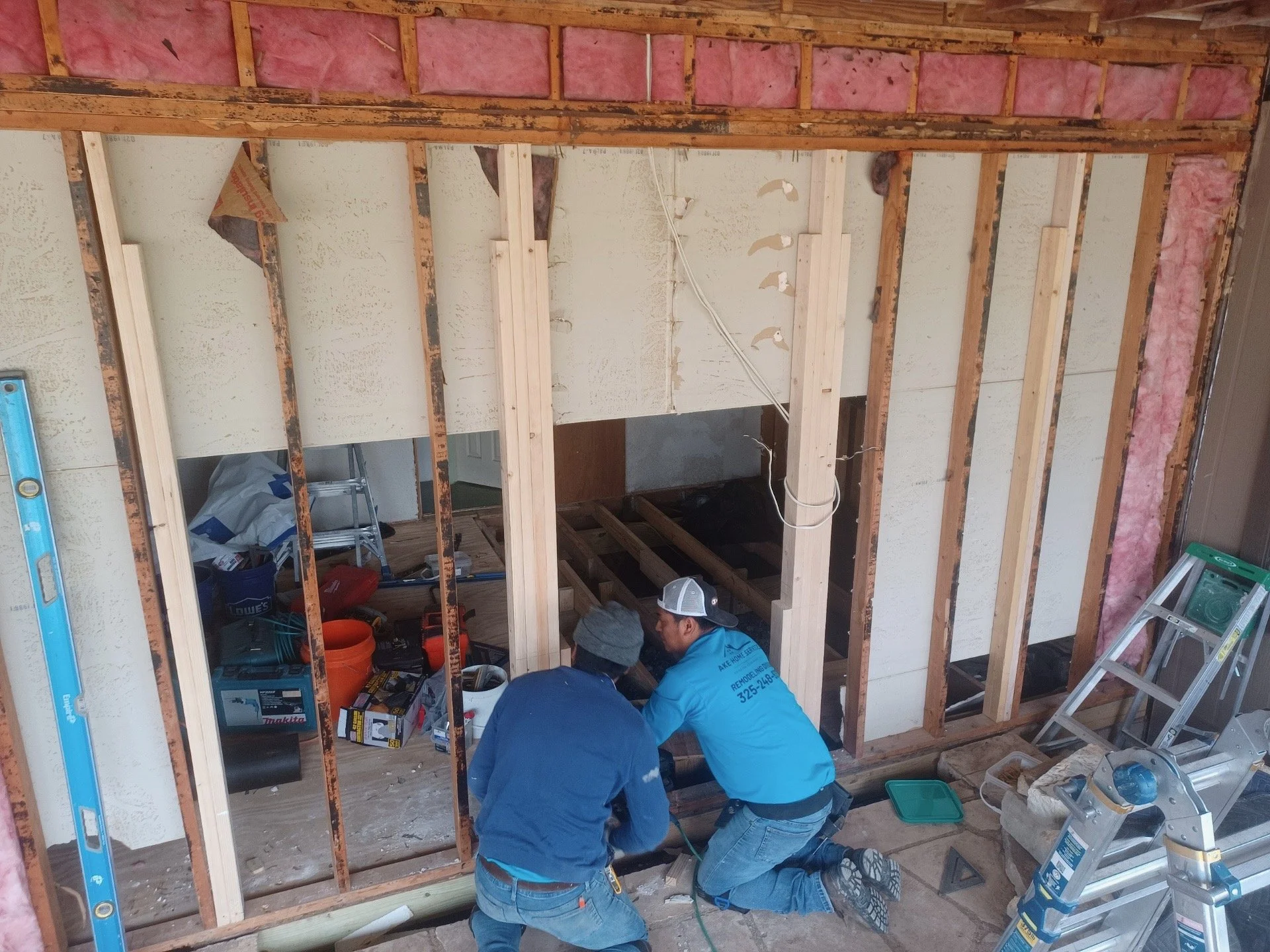 Two workers kneel on the floor and work on framing a wall inside a house under construction, surrounded by construction tools and equipment.