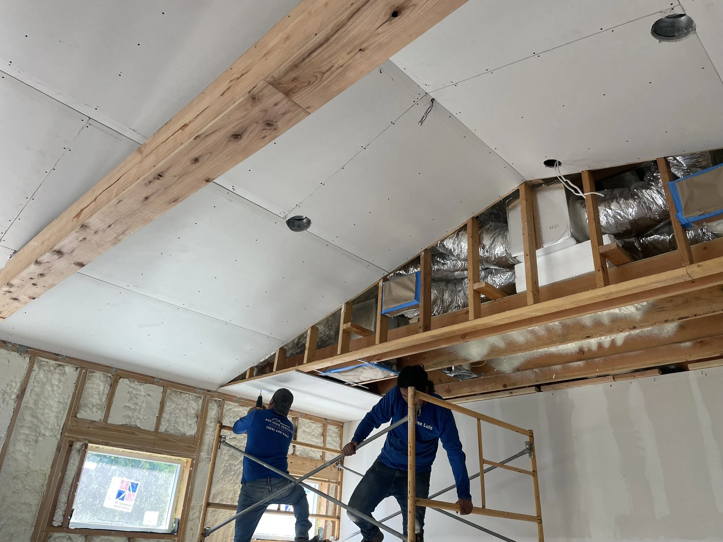 Construction workers installing drywall on the ceiling while standing on scaffolding inside a house under renovation.