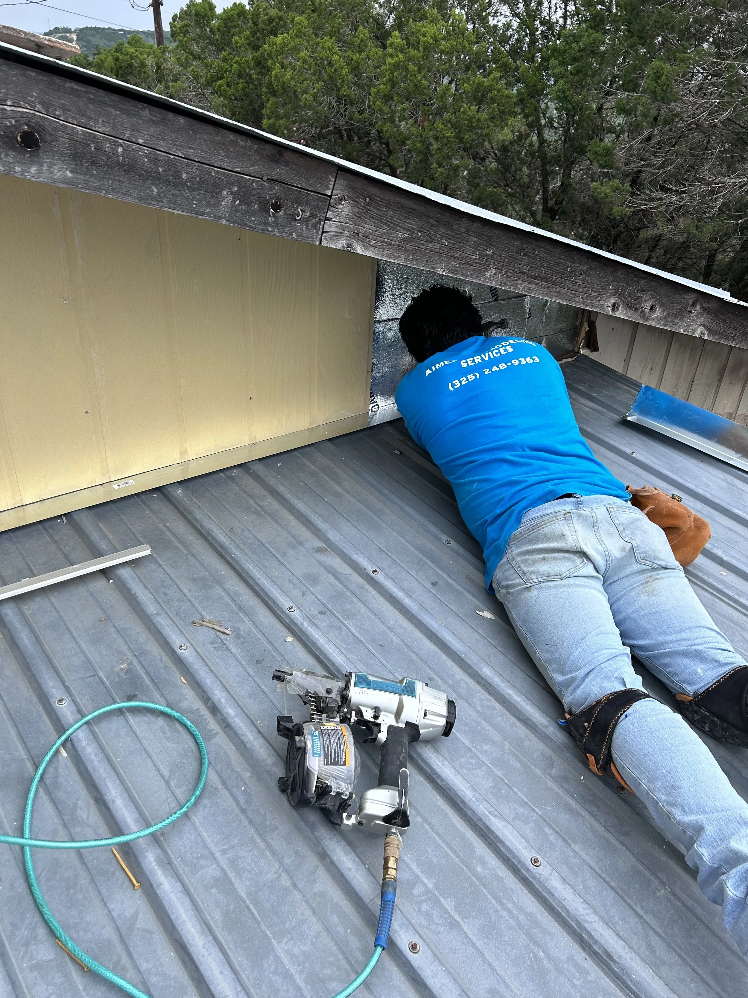 A worker installing or repairing metal roofing on a house with a cordless nail gun. The worker is lying on the metal roof, wearing a blue shirt and jeans, with trees and a hillside in the background.