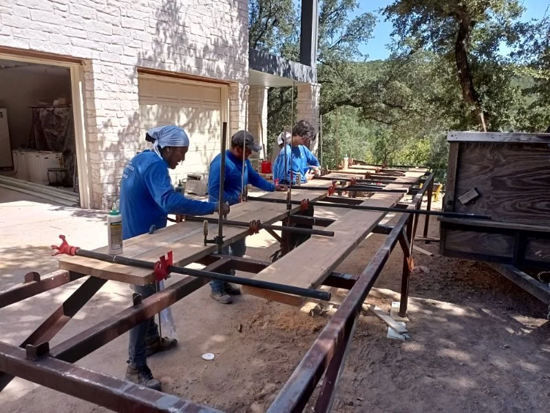 A group of five people working together on a woodworking project outdoors, using clamps to hold pieces of wood in place. They are dressed in blue shirts, with some wearing head coverings, and are positioned along a long workbench.